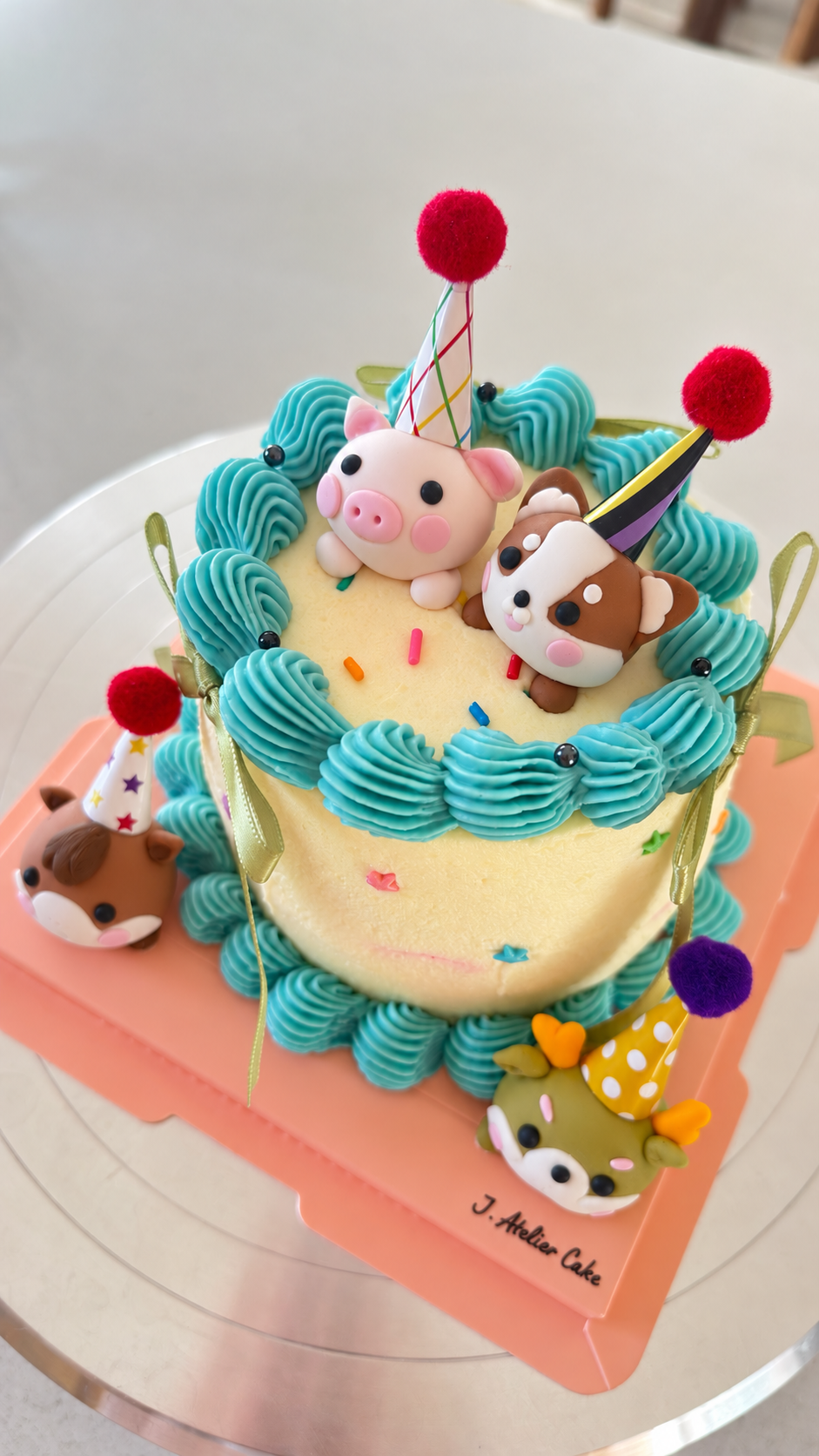Playful birthday cake with handmade fondant pig, dog, and deer characters wearing party hats, finished with bright blue piping, ribbons, and colorful sprinkles.