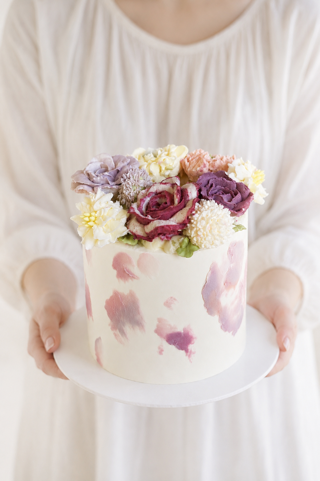 Person in a light-colored dress holding a round white luxury buttercream floral cake decorated with watercolor pink and purple flowers on top.