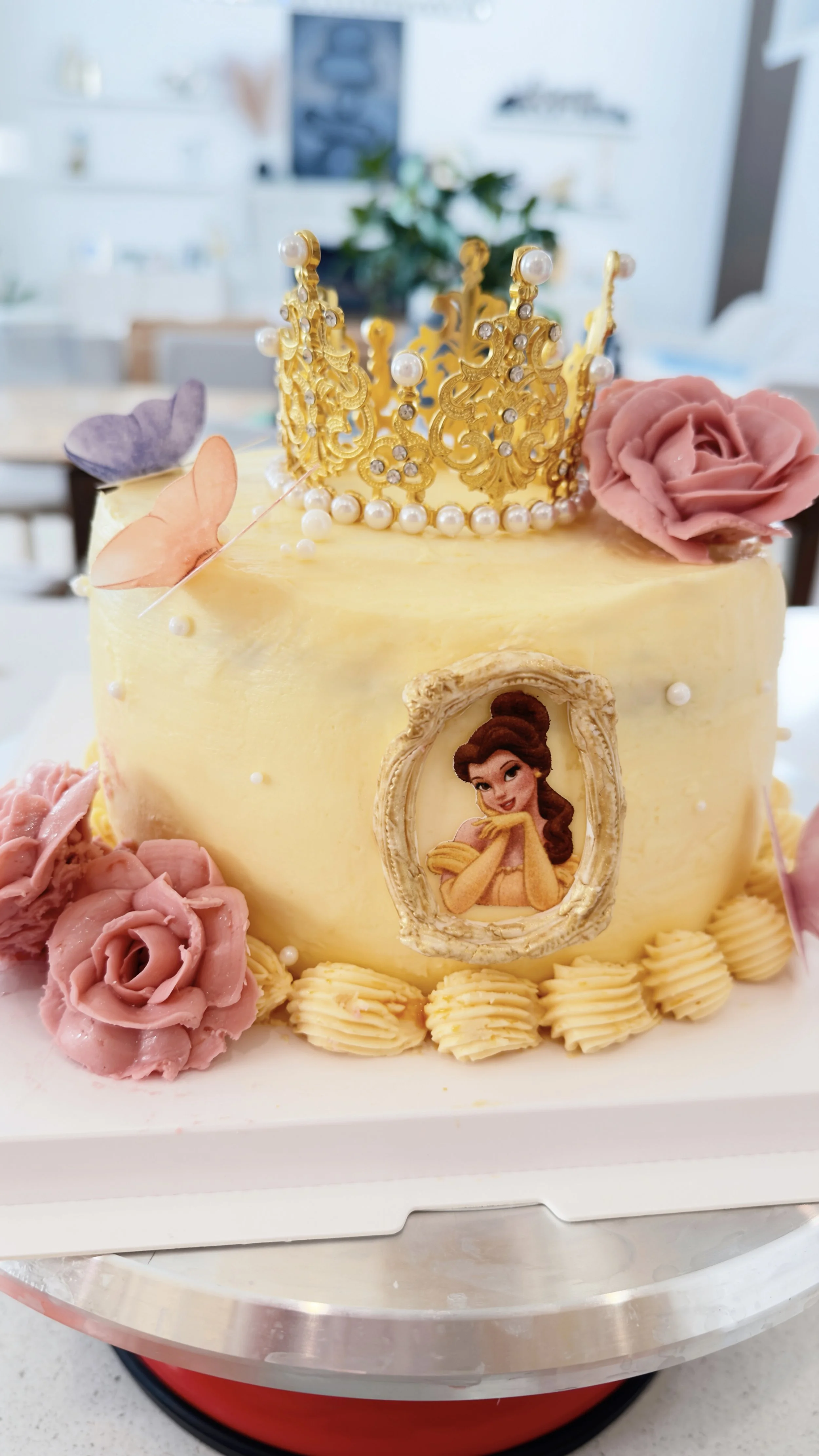 Elegant princess-inspired birthday cake with a gold crown, pink buttercream roses, delicate butterflies, pearls, and a Belle portrait.