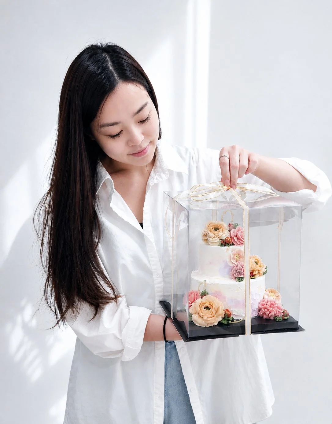 A woman holding a clear gift box containing a decorated wedding cake with pink and beige buttercream flowers, tied with a gold ribbon.