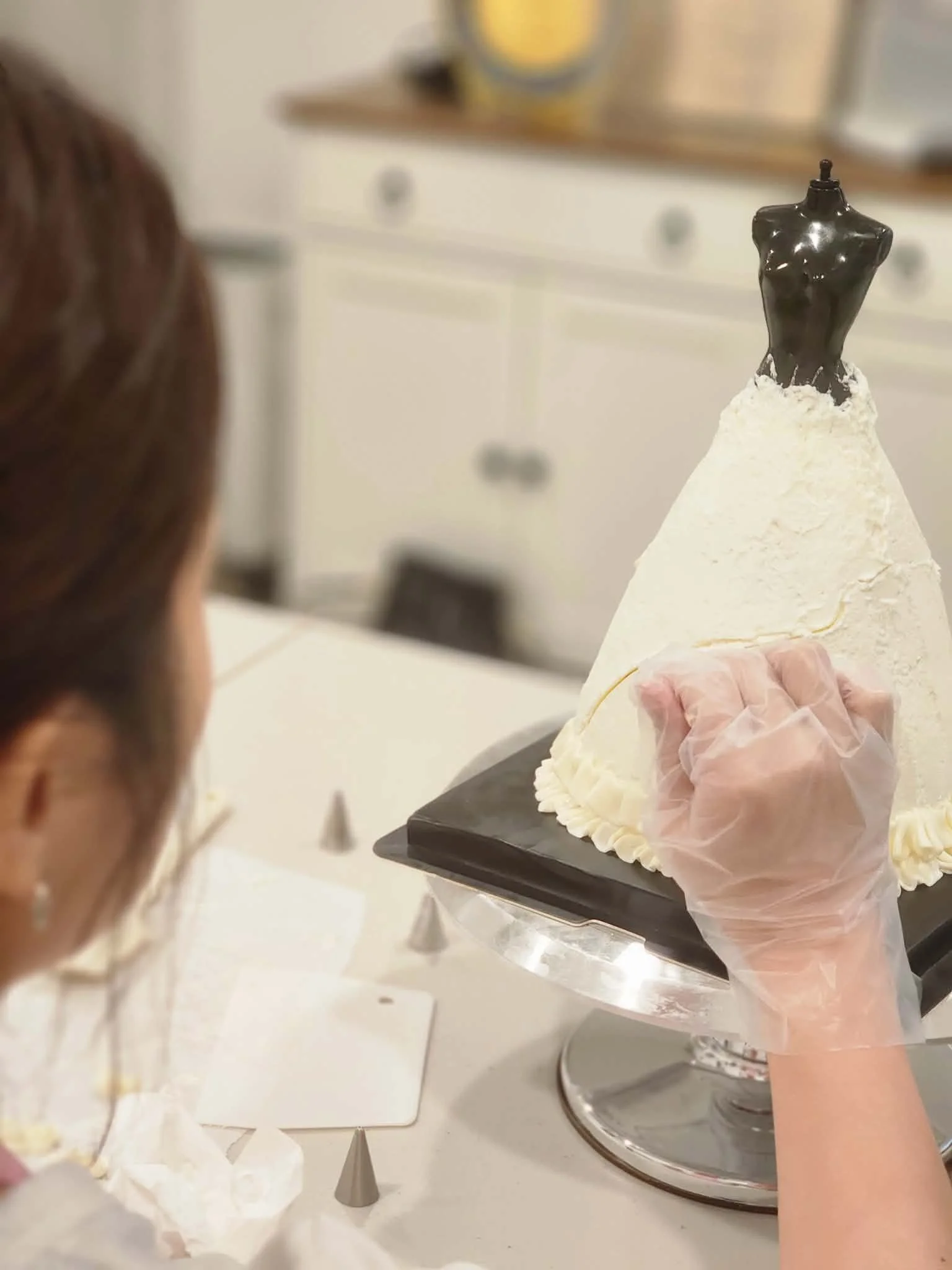 A person decorating a wedding dress cake with white frosting and a black mannequin topper at a bakery & kitchen counter.