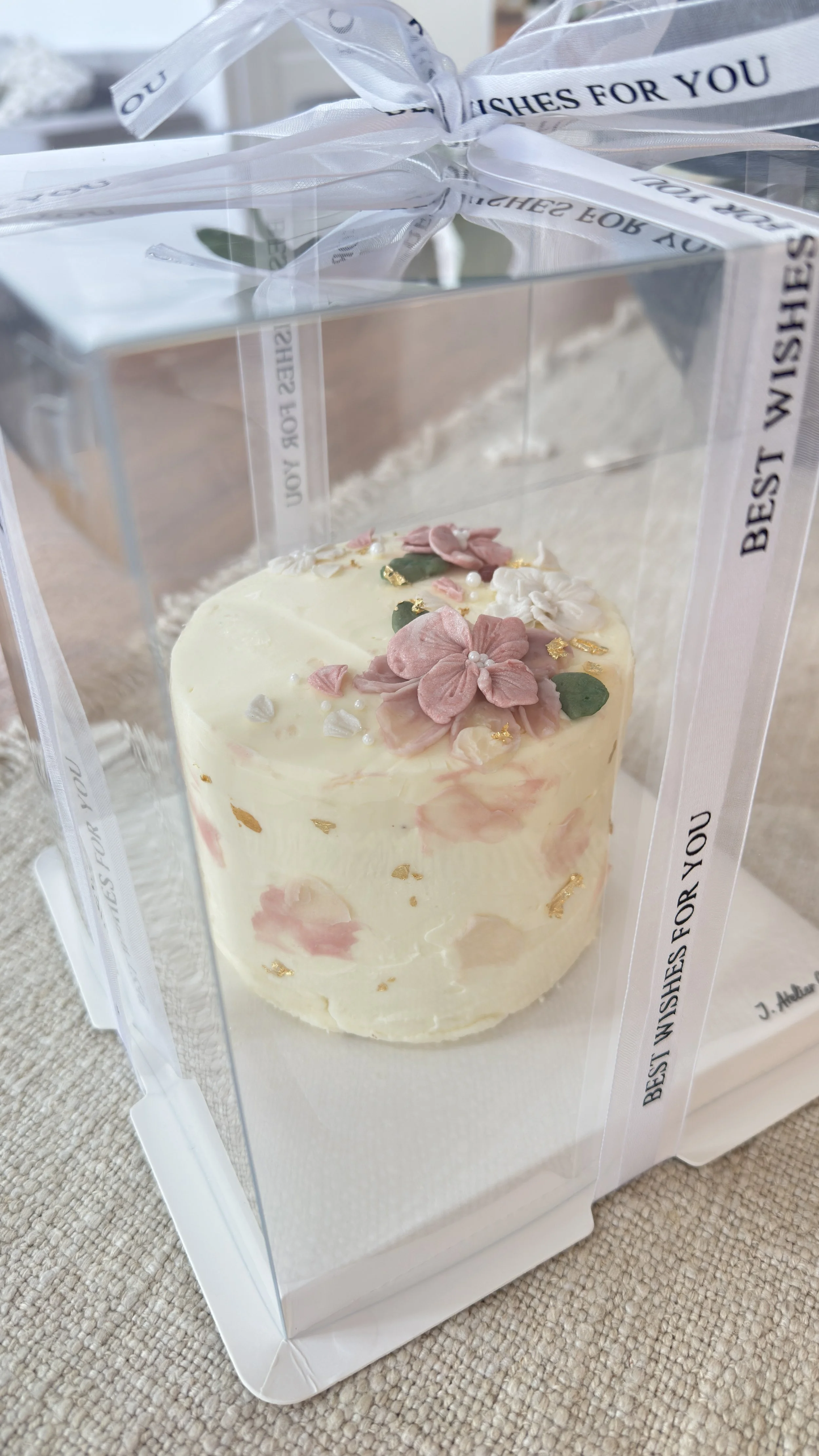 Elegant Korean-style floral cake with soft pink buttercream flowers, delicate gold accents, and green leaves, presented in a clear luxury gift box.