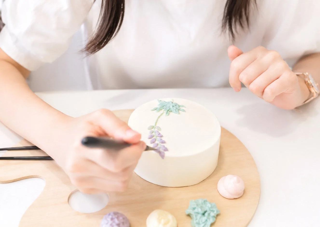 A person decorating a white cake with pastel-colored floral icing accents with a black decorating tool, on a round wooden board, with small decorative meringue-like balls nearby. DIY kit includes everything.