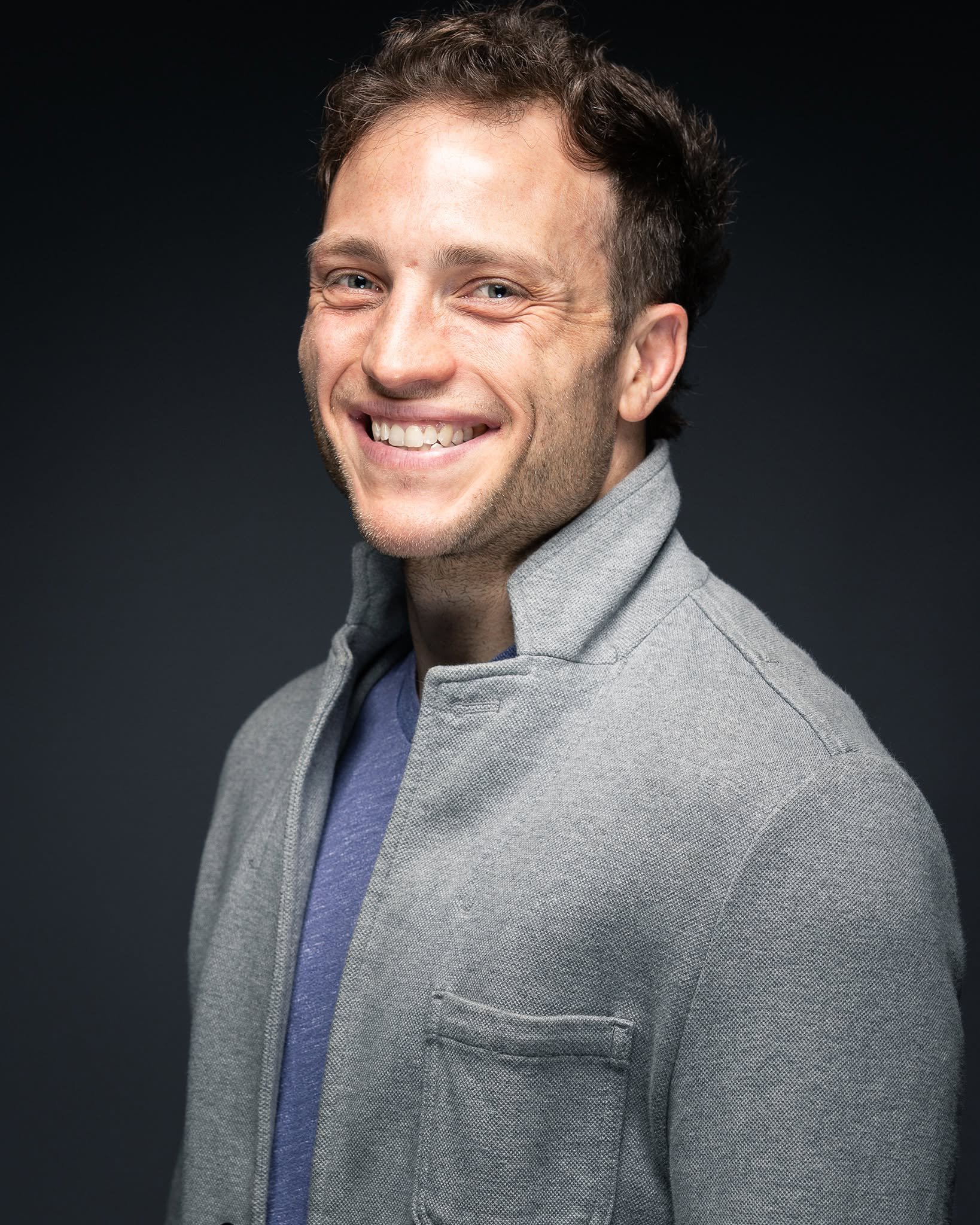 Close-up of a smiling man wearing a gray blazer and a blue shirt, against a dark background.