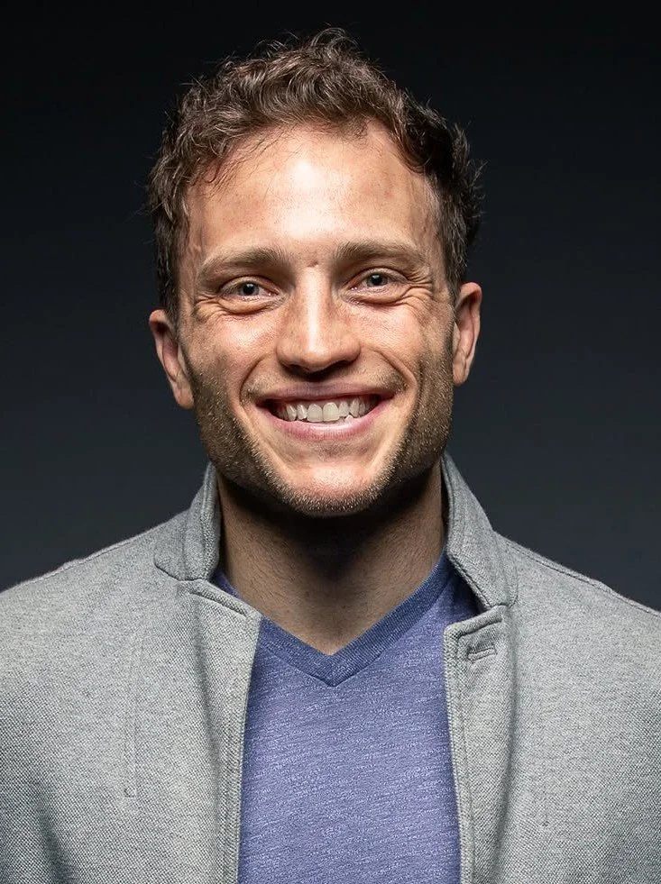 A young man with light skin, curly light brown hair, and blue eyes smiling at the camera. He's wearing a gray jacket over a blue V-neck shirt, with a dark background.