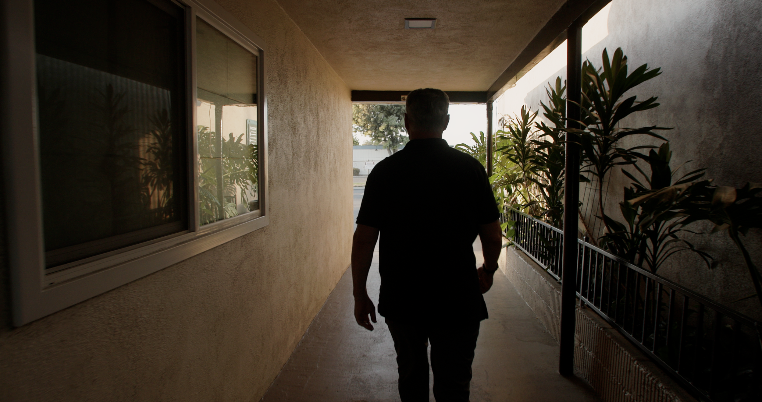A man walking through an outdoor corridor with beige walls and a row of green plants on the right side.