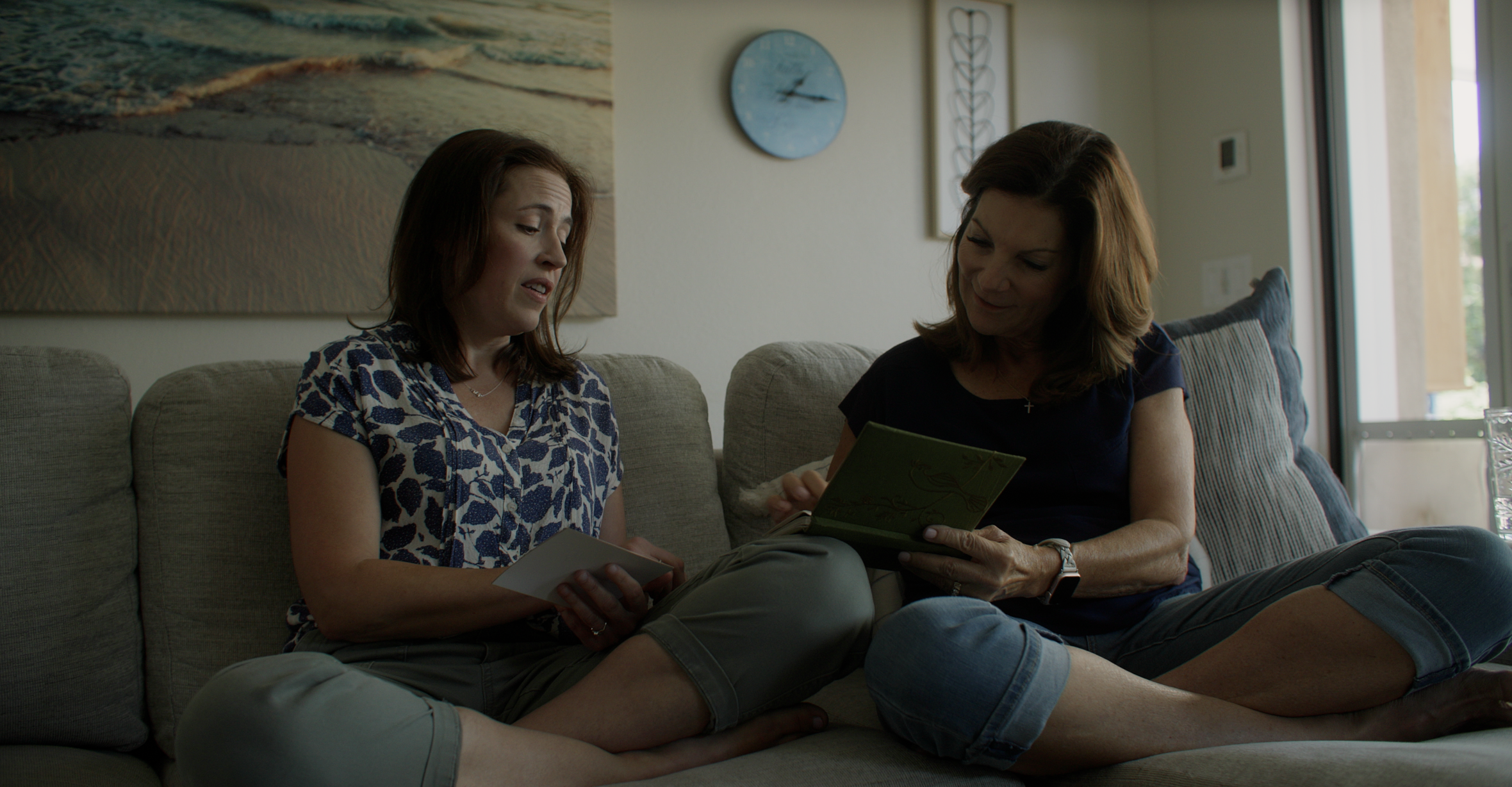 Two women sitting on a beige couch indoors, engaged in reading a book together, with a window and wall decorations in the background.