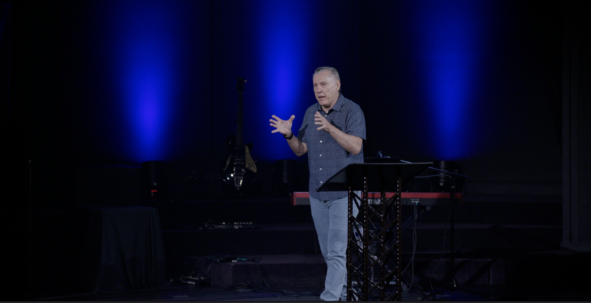 A man with short gray hair wearing a patterned shirt and light-colored jeans is speaking on a stage with blue lit background. There is a music stand and a red keyboard on stage, with guitars visible in the background.