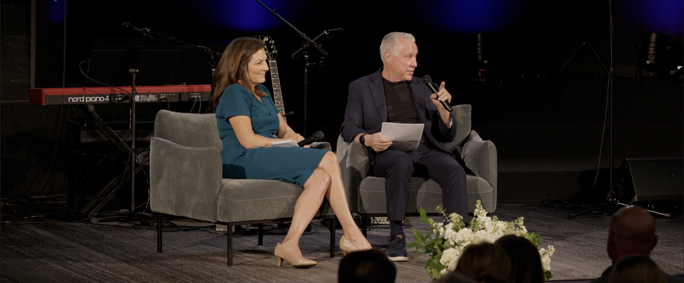 Two people sitting on a stage during a panel discussion or interview, with musical instruments and equipment behind them, and audience members visible at the bottom of the image.