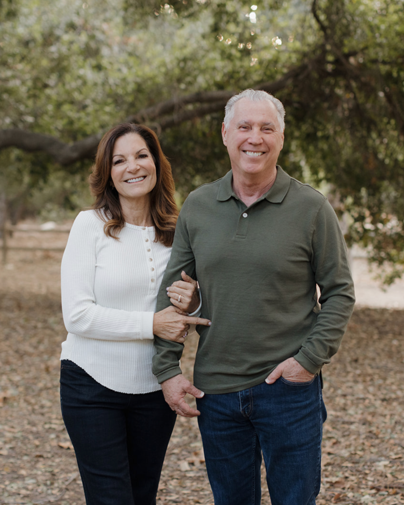 A smiling middle-aged couple standing outdoors in a park with green trees in the background. The woman, with long brown hair, wearing a white ribbed sweater, is holding the arm of the man who has short gray hair and is wearing a green long-sleeve polo shirt. They are standing close together, smiling at the camera.