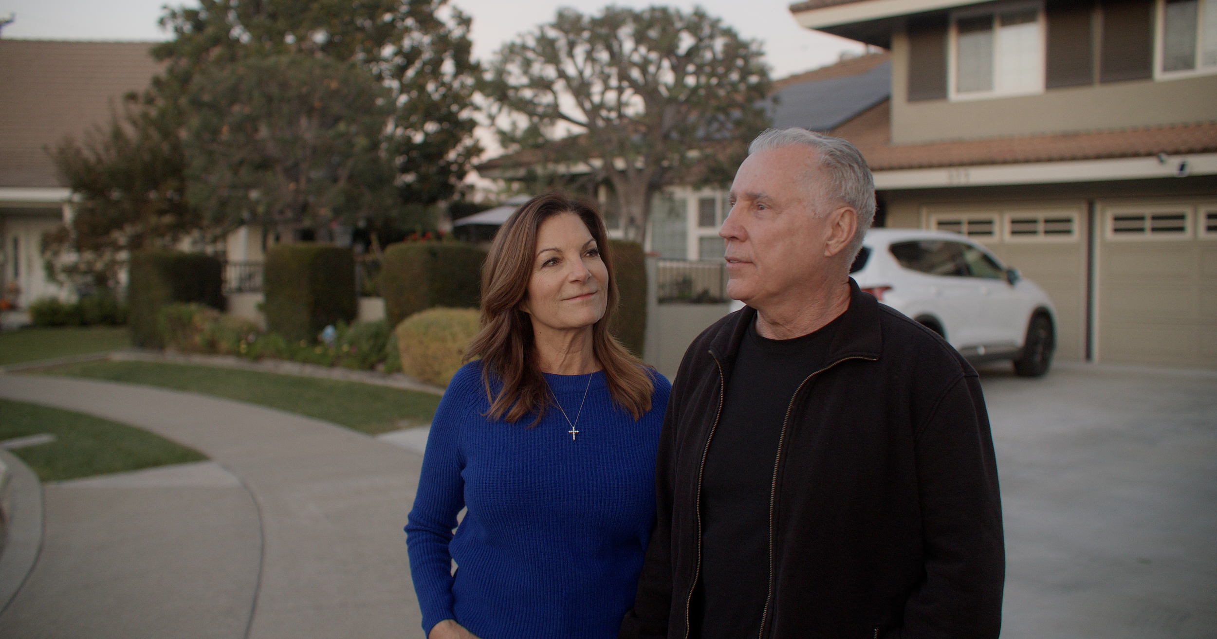 An older man and woman standing outside in a residential neighborhood, facing each other with the woman looking at the man. The woman has long brown hair and is wearing a blue sweater with a cross necklace. The man has short gray hair and is wearing a black jacket. There are houses, trees, and a white vehicle in the background.