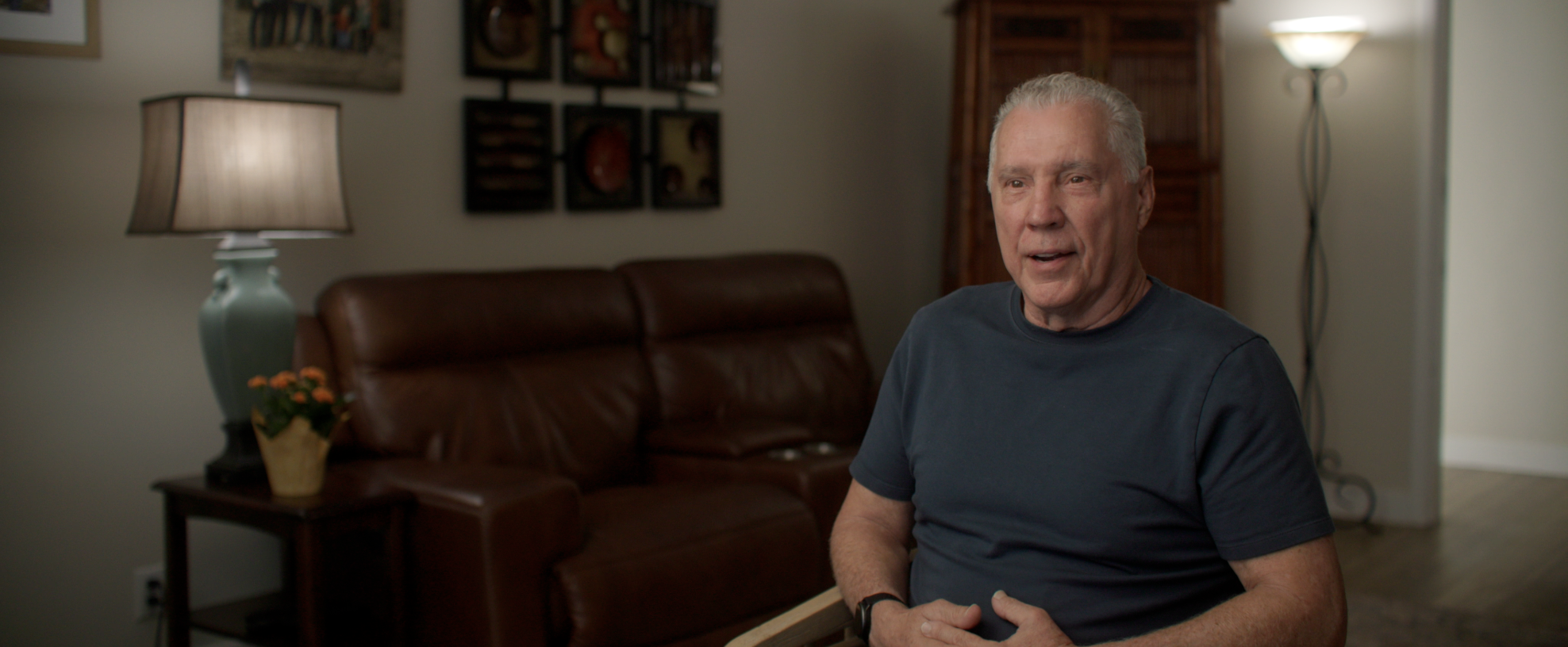 An elderly man sitting and talking in a living room with a brown leather couch, a lamp, and wall art in the background.