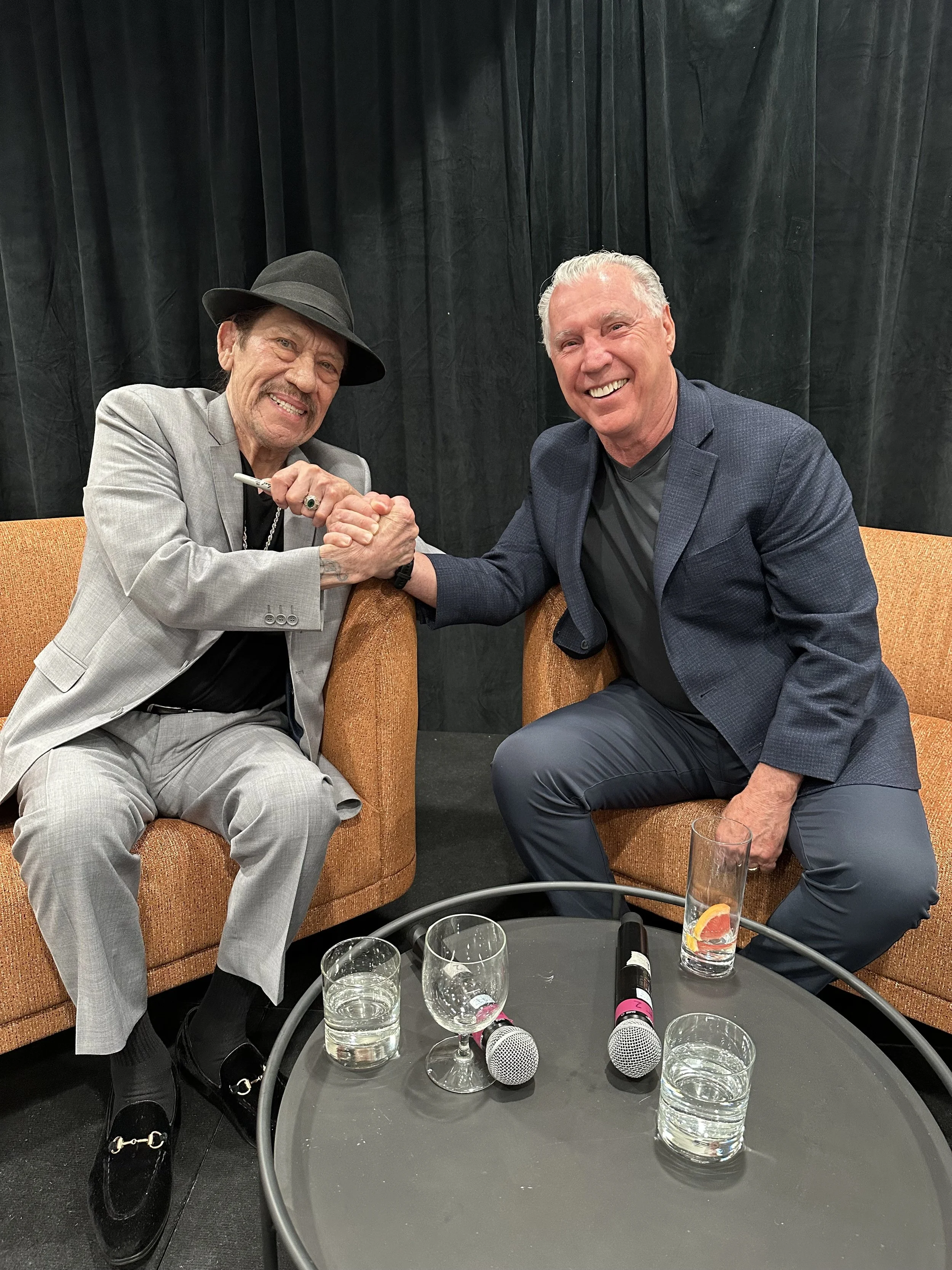Two men sitting on orange couches shaking hands and smiling at the camera. The man on the left is older, wearing a grey suit and black hat, and holding a marker. The man on the right is younger with grey hair, wearing a dark blue blazer and black shirt. In front of them are glasses of water, microphones, and a pink marker on a black round table, with a black curtain backdrop.