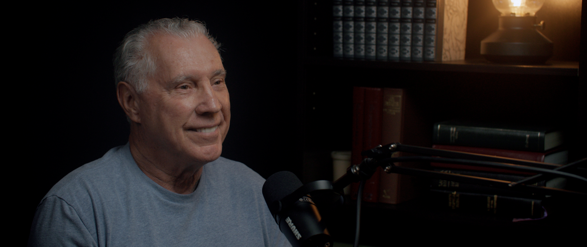 An elderly man with gray hair wearing a light blue shirt, smiling while speaking into a professional microphone in a dimly lit room with bookshelves and books in the background.