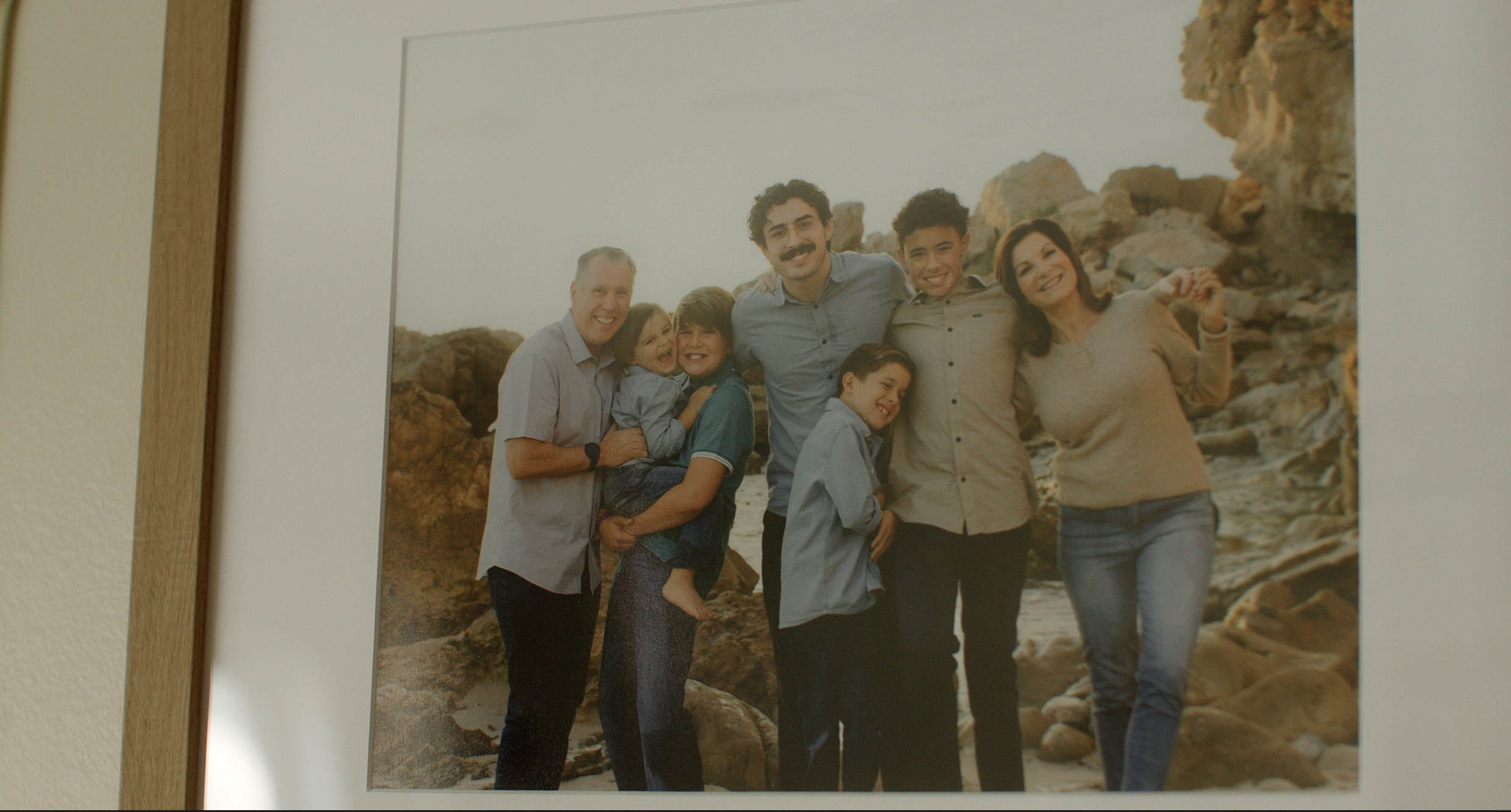 Group of seven people, including children, adults, and a pregnant woman, smiling and posing together on a rocky beach with large rocks in the background.