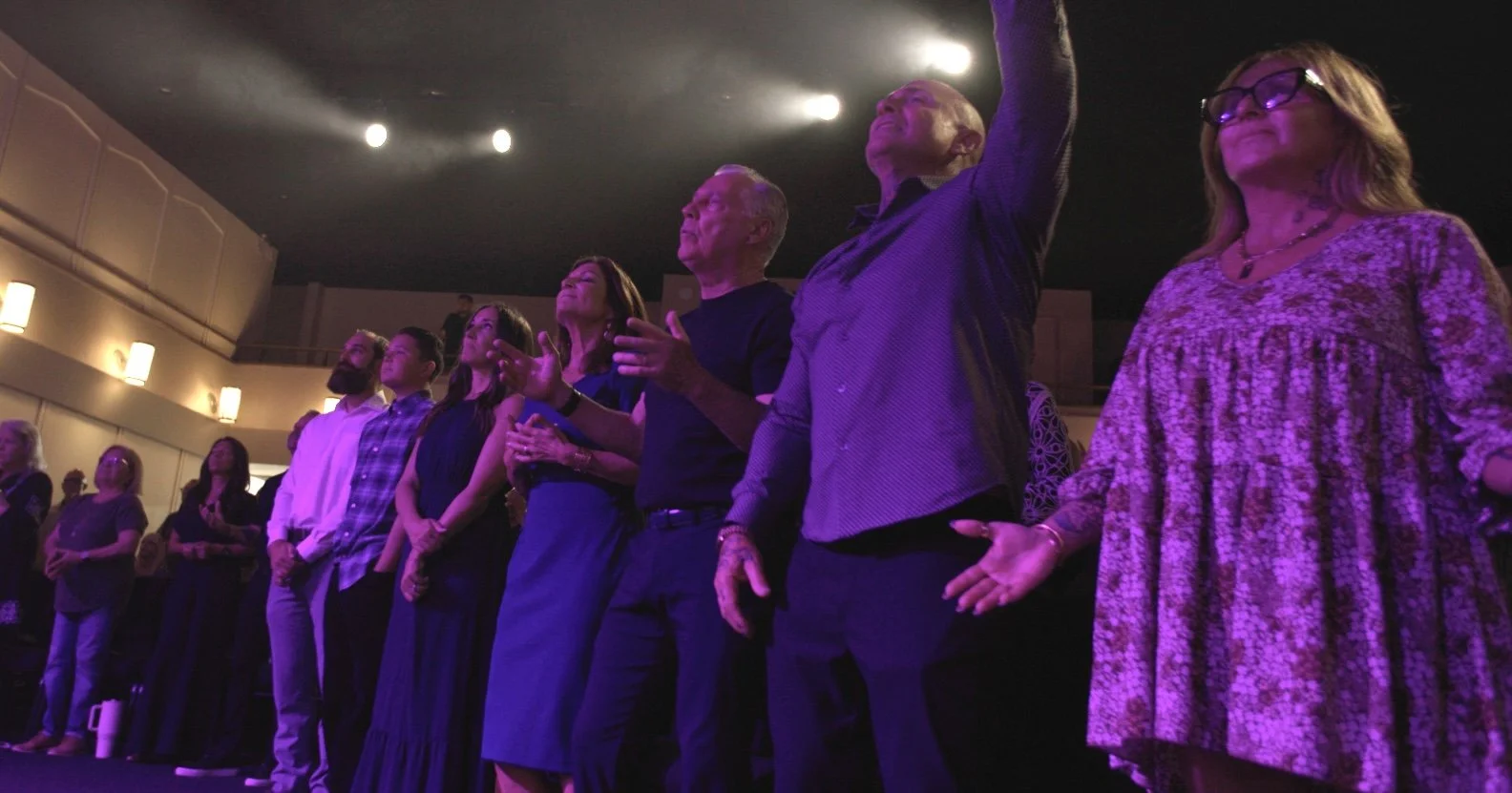 A diverse group of people standing on stage, some with eyes closed and hands raised, appears to be engaged in worship or prayer during a religious or spiritual event in a darkened auditorium with purple lighting.