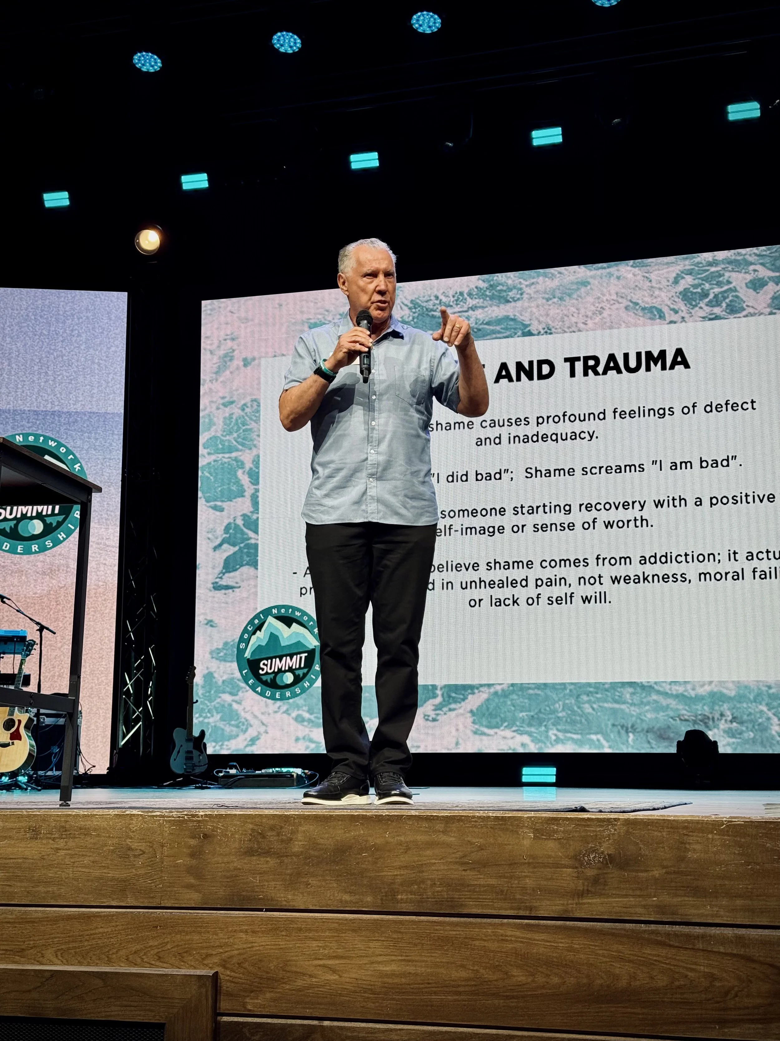 A man speaking on stage with a microphone in hand, standing in front of a presentation screen that reads 'Healing and Trauma' and discusses shame and recovery. The stage has guitars and music equipment, with blue lights above.
