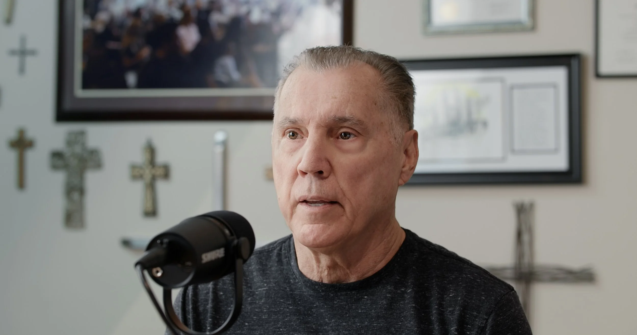 A middle-aged man with short gray hair speaking into a microphone while sitting in front of framed artwork and crosses on the wall.