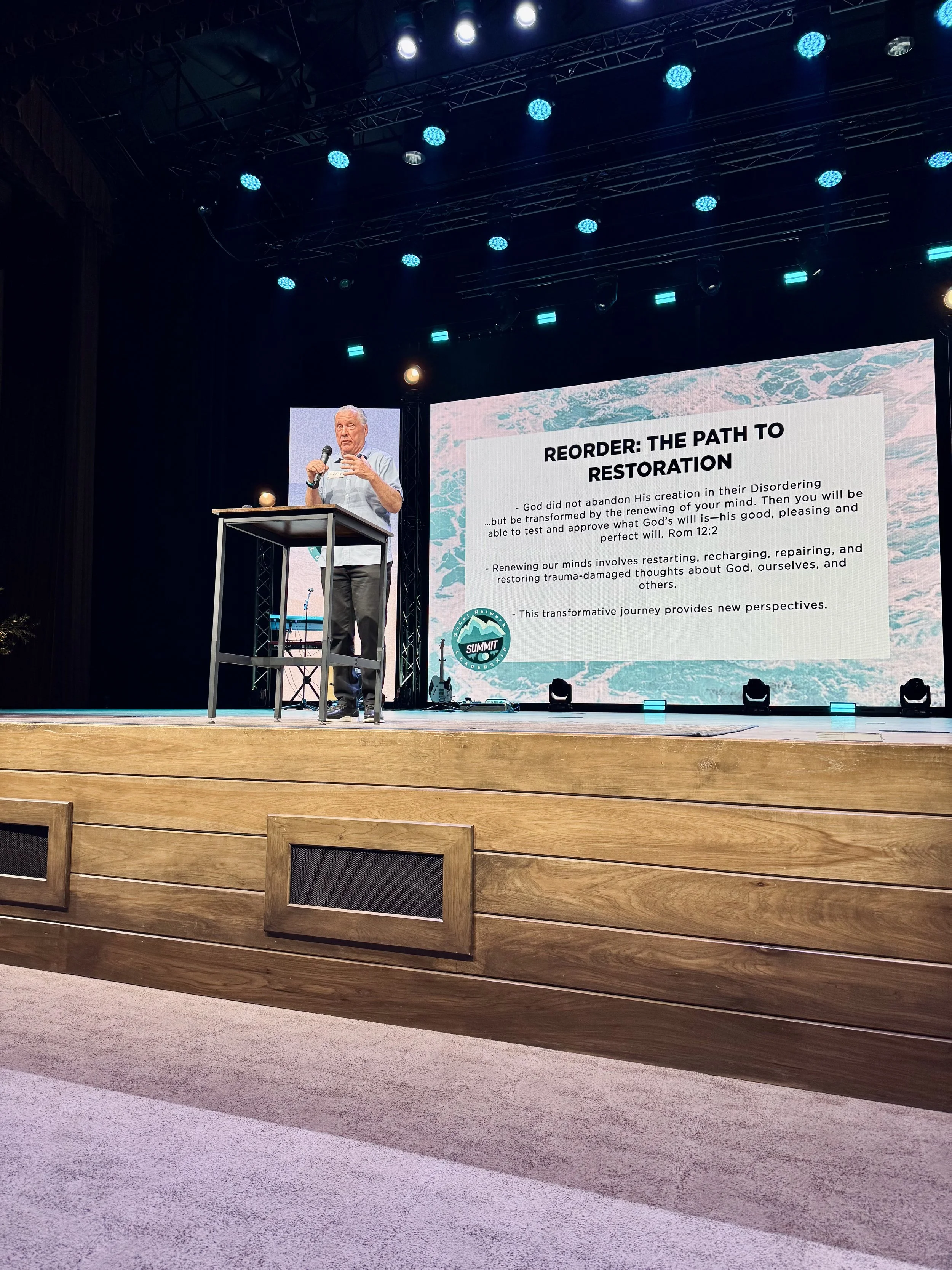 A man giving a presentation on a stage with a large screen behind him displaying a slide titled "Reorder: The Path to Restoration". The stage has bright overhead lights and a wooden front panel.