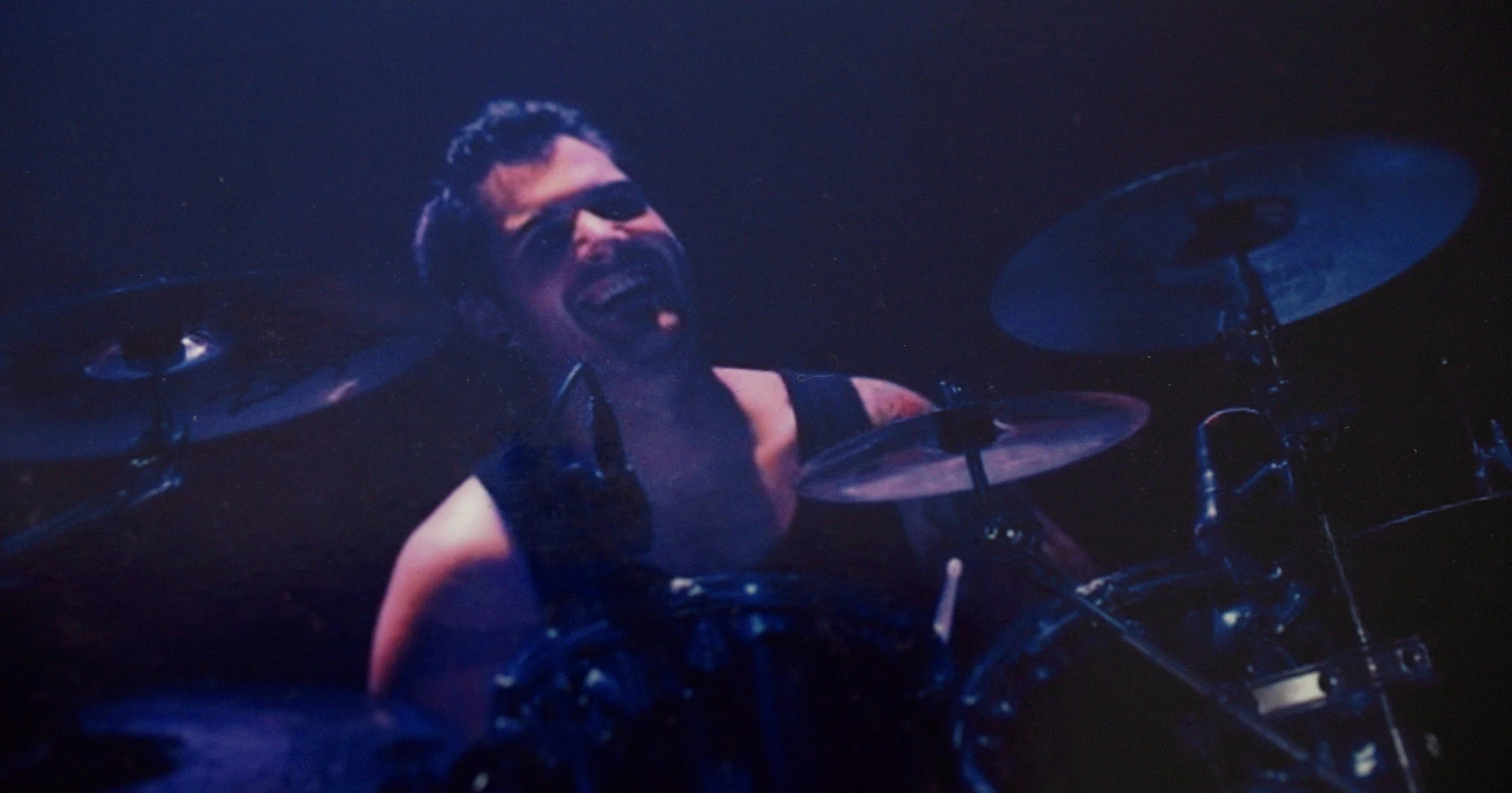 A drummer wearing sunglasses, a sleeveless shirt, and a beard, playing a drum set on stage in a dark environment.