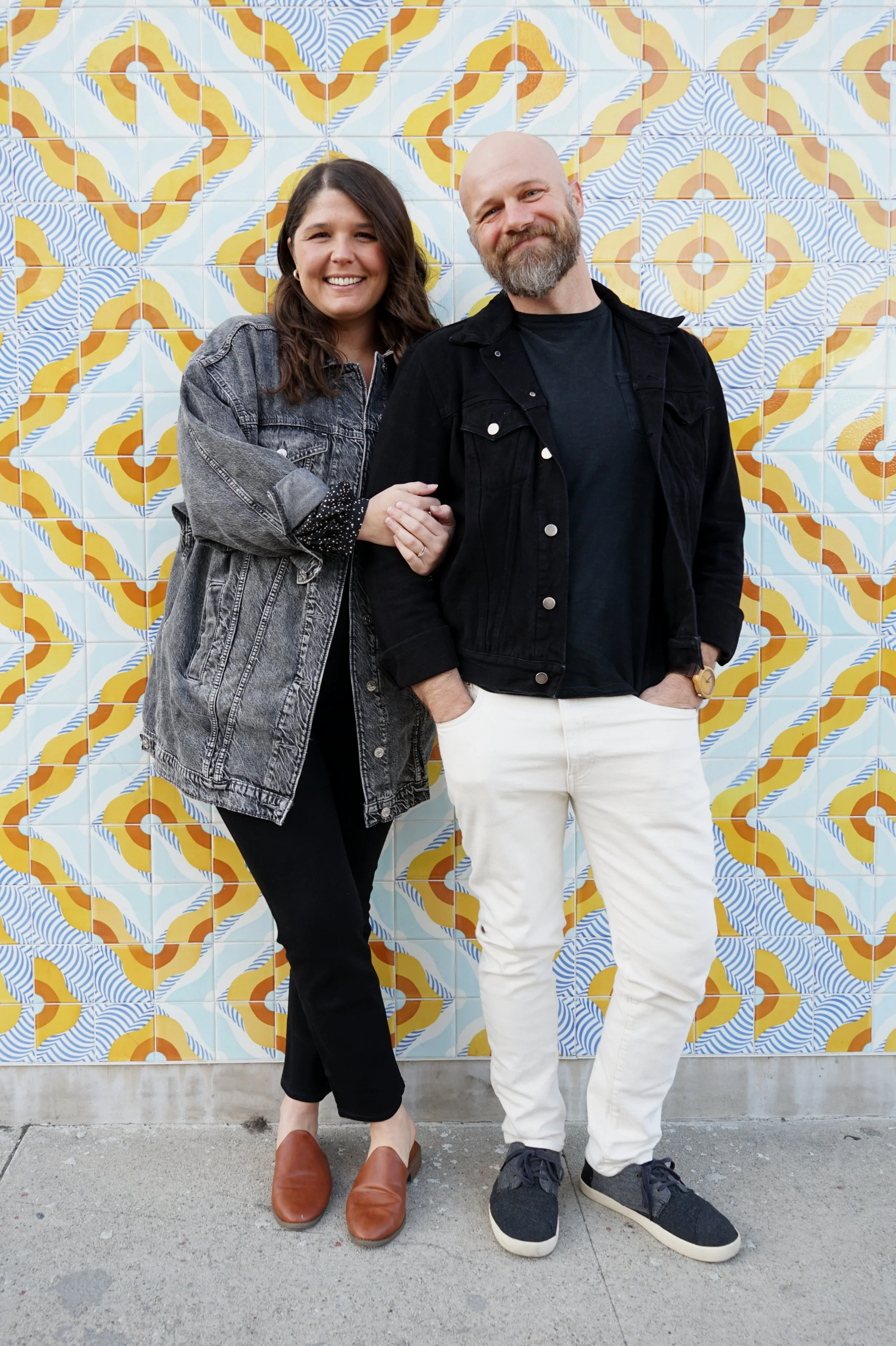 A smiling woman with brown hair wearing a denim jacket stands next to a smiling man with a beard wearing a black jacket, black shirt, and white pants. They are standing in front of a colorful patterned tile wall.