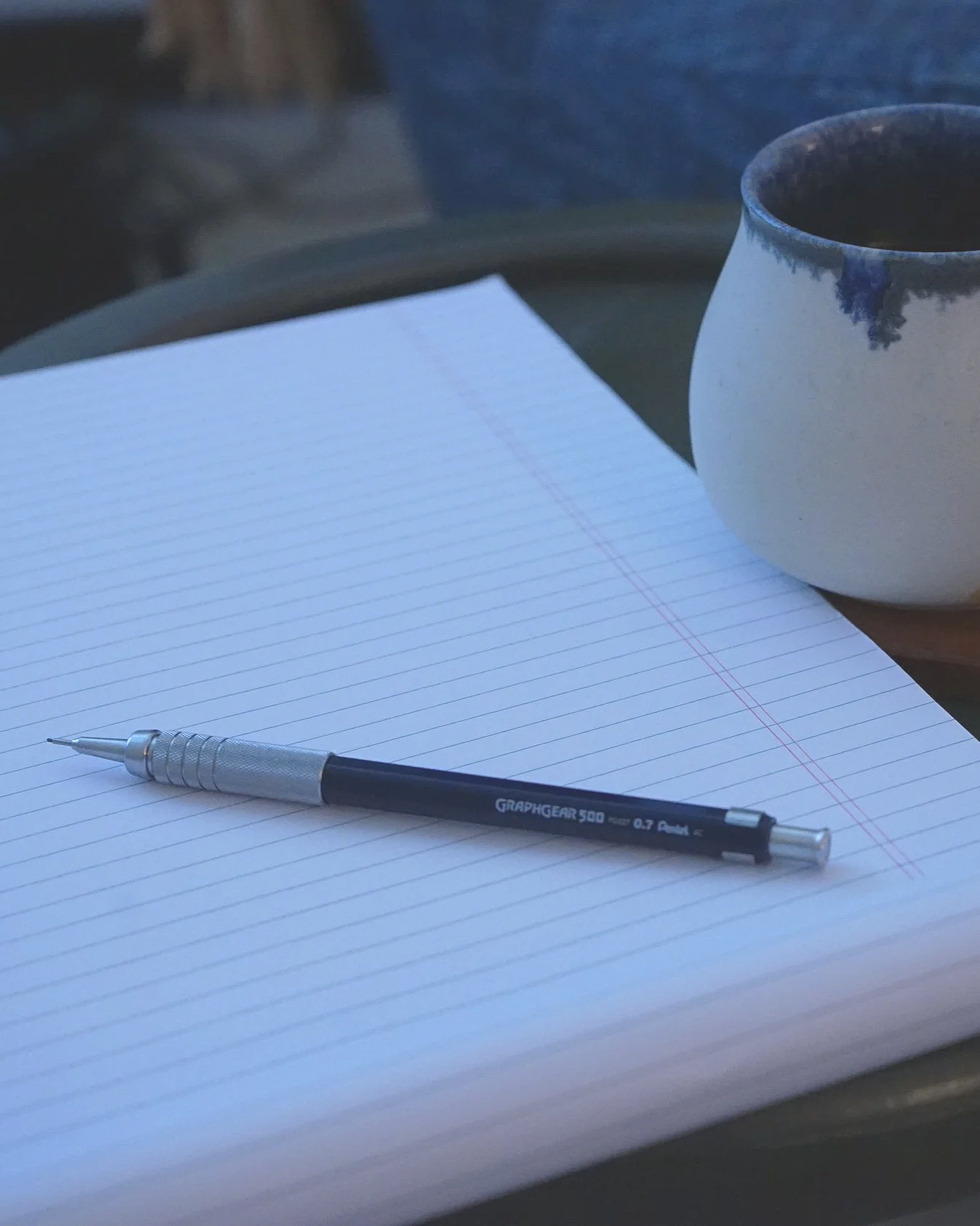A notebook with lined paper and a mechanical pencil resting on it, a white mug, and a blue and white ceramic cup on a table.