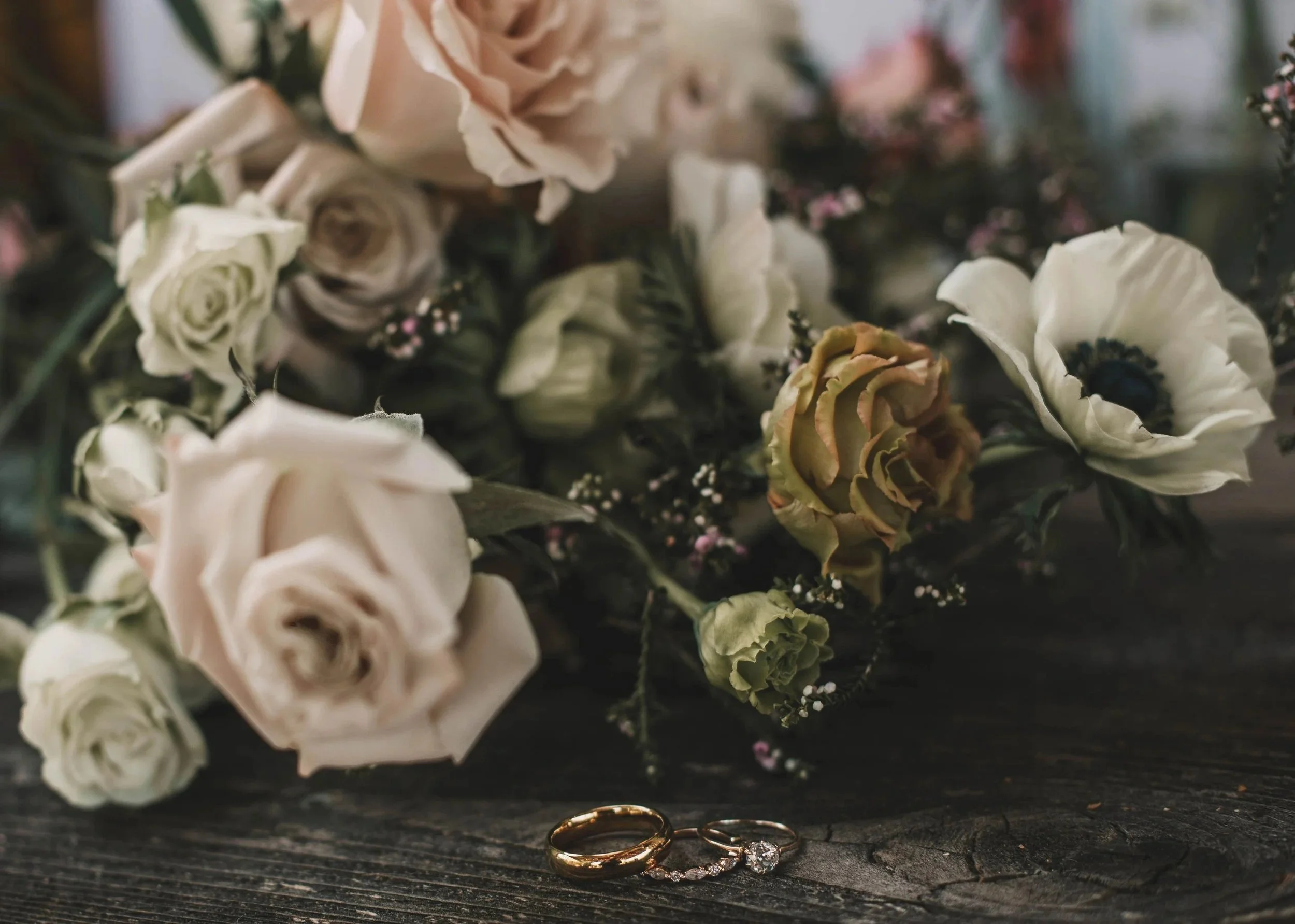 Close-up of wedding rings, including a gold band and a diamond ring, on a rustic wooden surface with a floral bouquet in the background.
