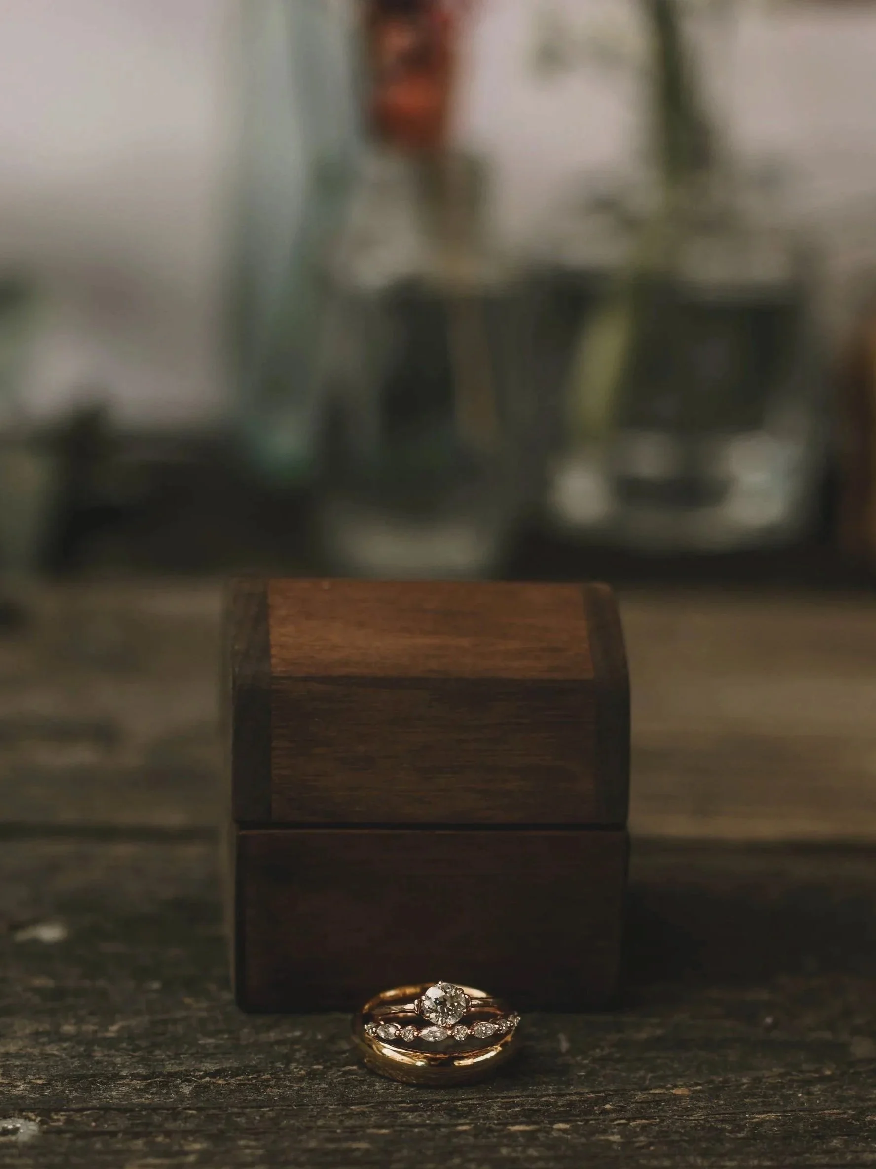 A pair of gold rings with diamonds, one with a large center diamond and smaller diamonds around it, placed on a dark surface in front of a glass case with rings inside.