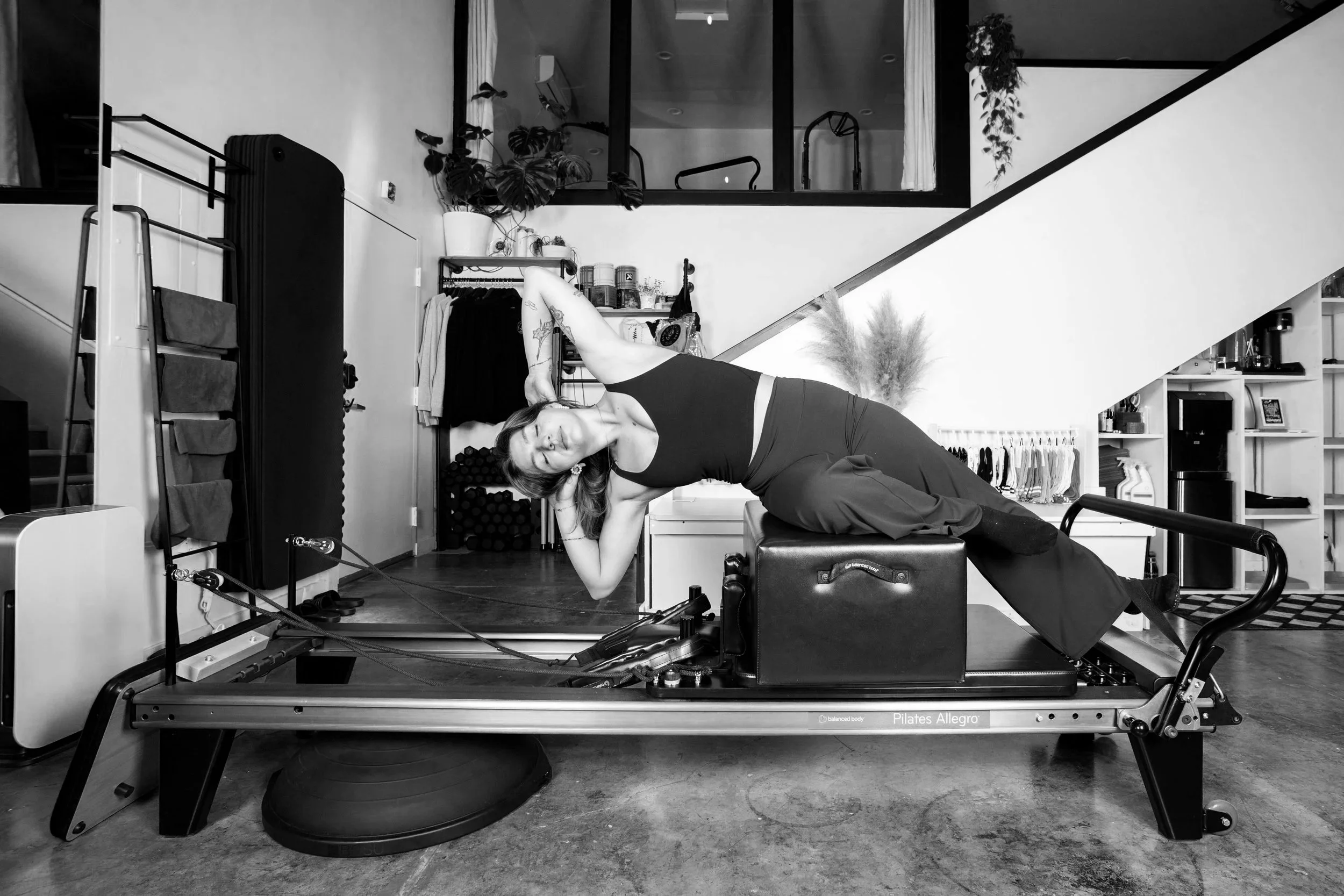 A woman practicing Pilates on a reformer machine in a modern indoor space with plants and storage shelves.