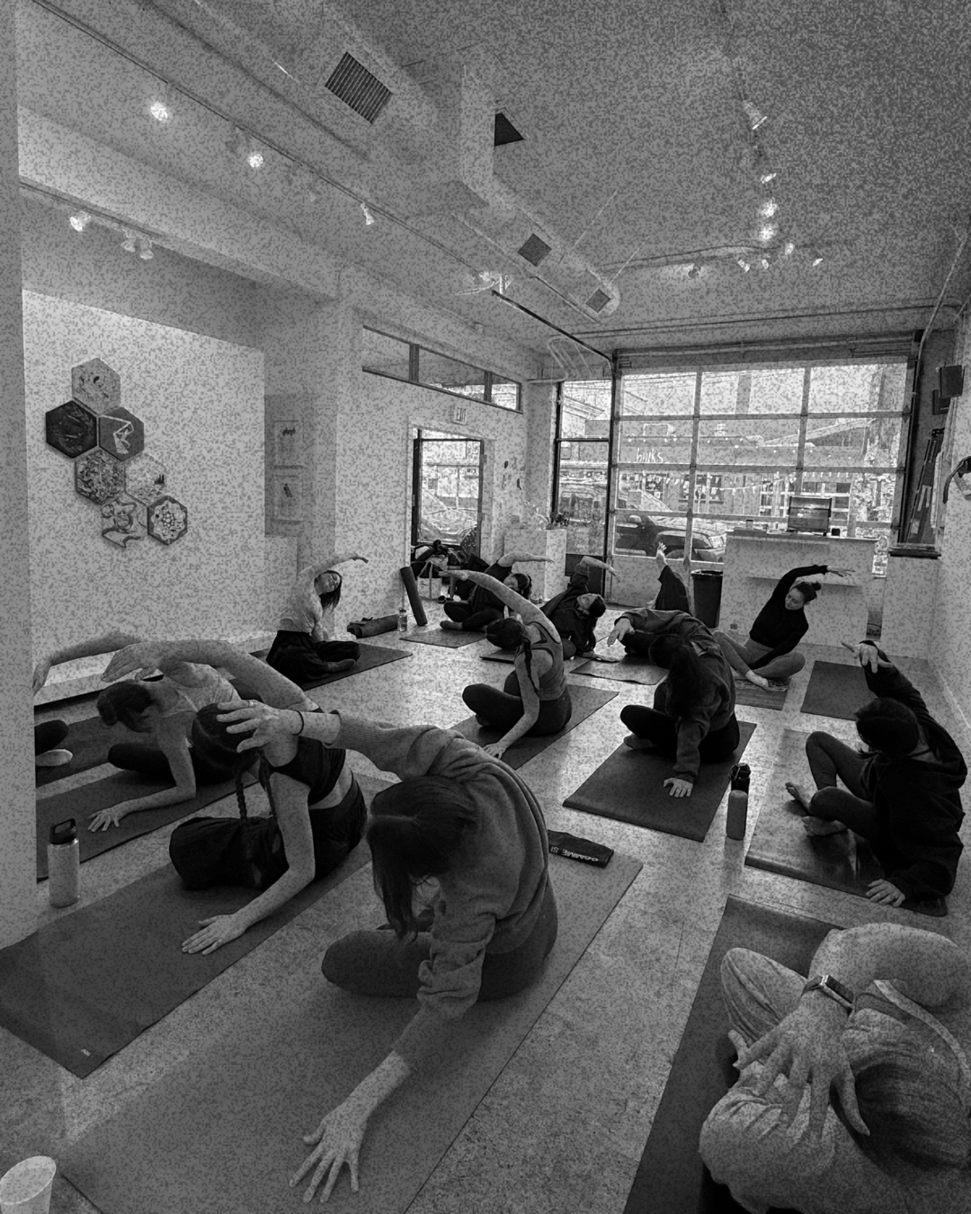 Group of people practicing yoga in a studio, sitting on mats, stretching with arms overhead, in black and white.
