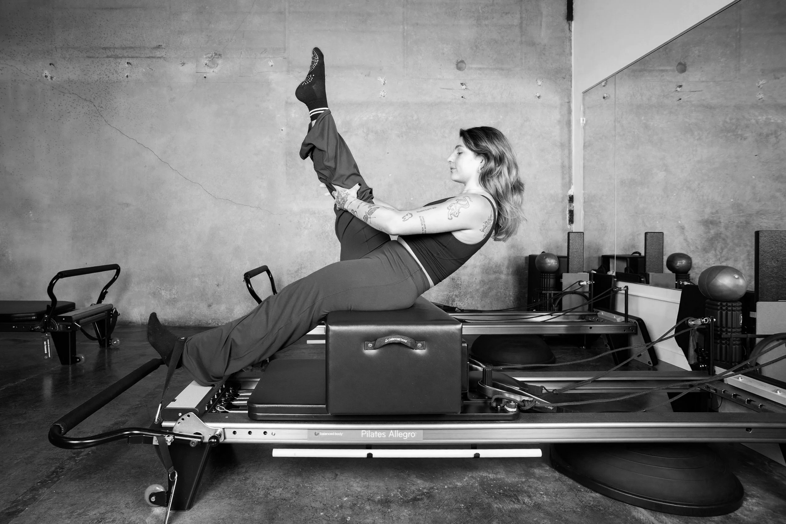 A woman practicing Pilates on a reformer machine in a gym, with a concrete wall background.