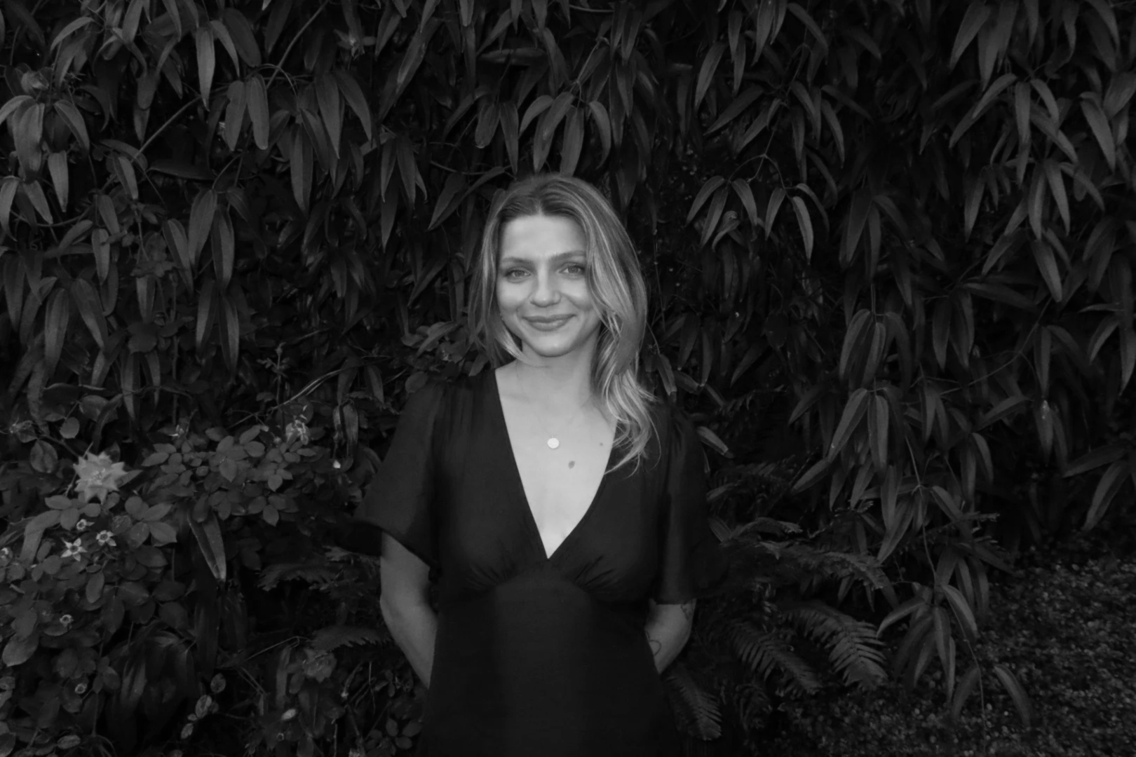 A woman standing in front of a leafy hedge, smiling, in a black dress, in black and white.