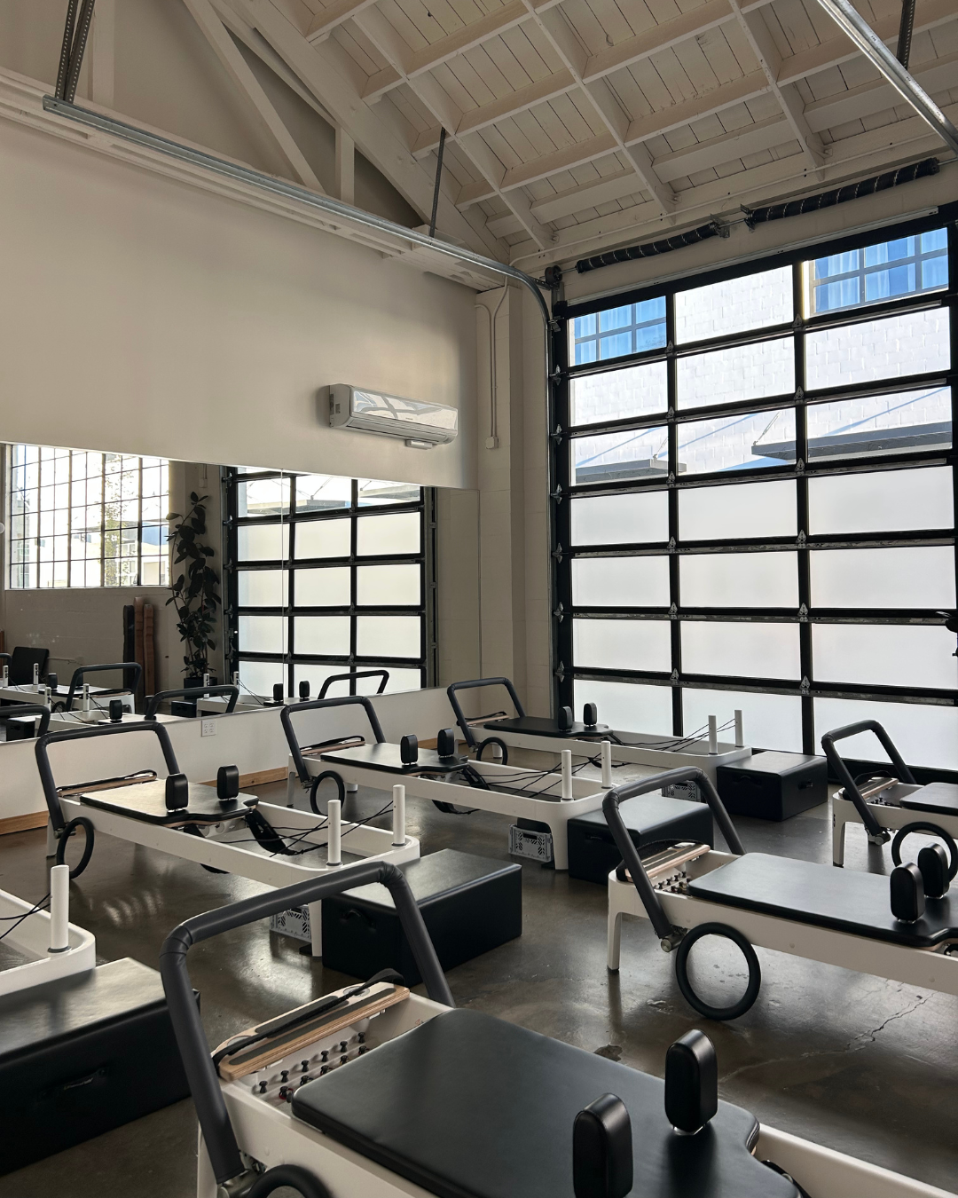 A Pilates studio with reformer machines arranged in rows, large windows with frosted glass, a wall mirror, and minimal decor.