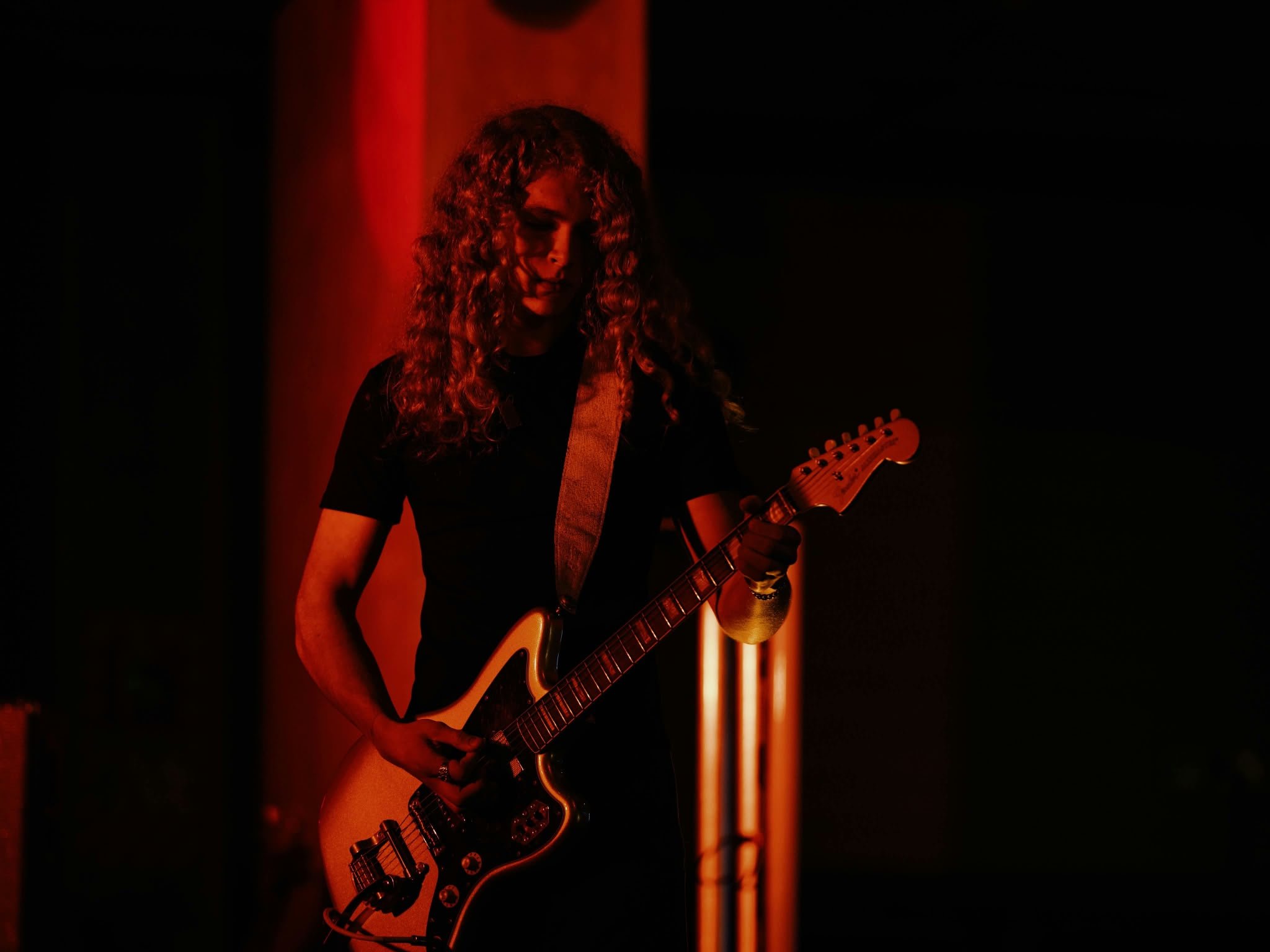 A person with long curly hair playing an electric guitar in a dimly lit environment with red lighting.