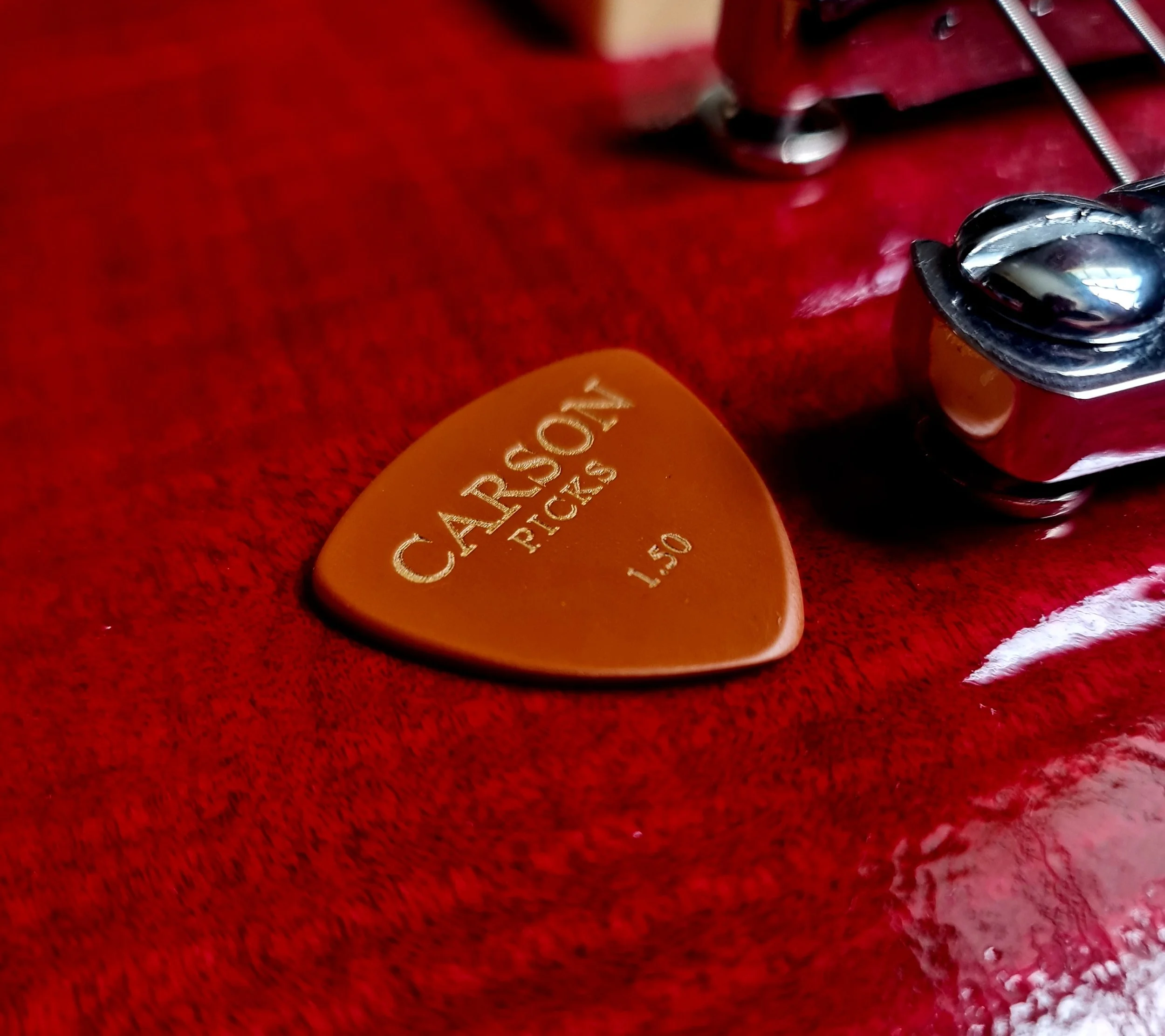 Close-up of a guitar pick made of Vespel SP-1 by Carson Picks, with "CARSON PICKS 1.50MM" engraved, lying on a red surface, next to a part of an electric guitar with strings and a metallic tuning peg visible. Les Paul