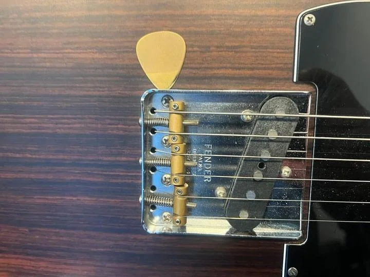 Close-up of a Fender electric guitar body showing the bridge, strings, and a Carson Picks brass custom pick tucked underneath the bridge with a shiny tip and textrued grip area.