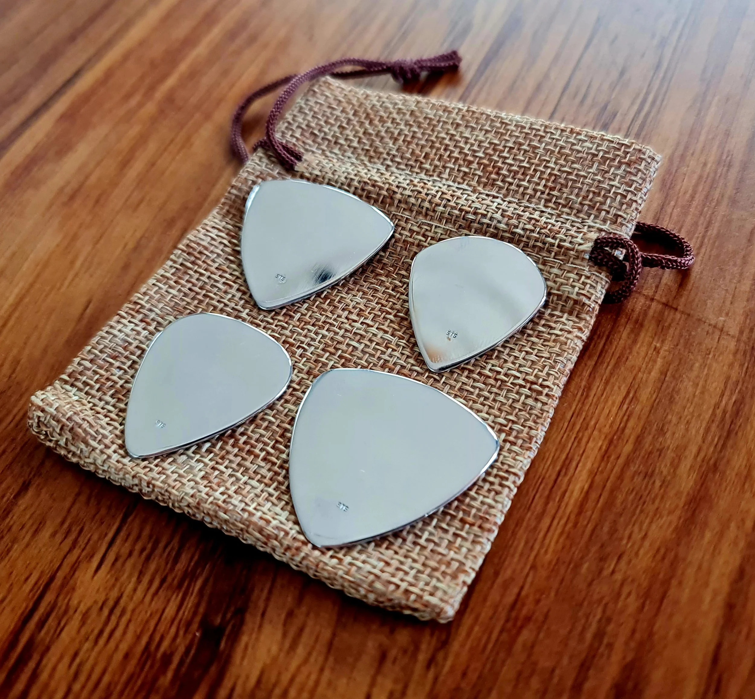 4 Sterling Silver Carson guitar picks resting on a woven, beige drawstring pouch on a wooden surface.
