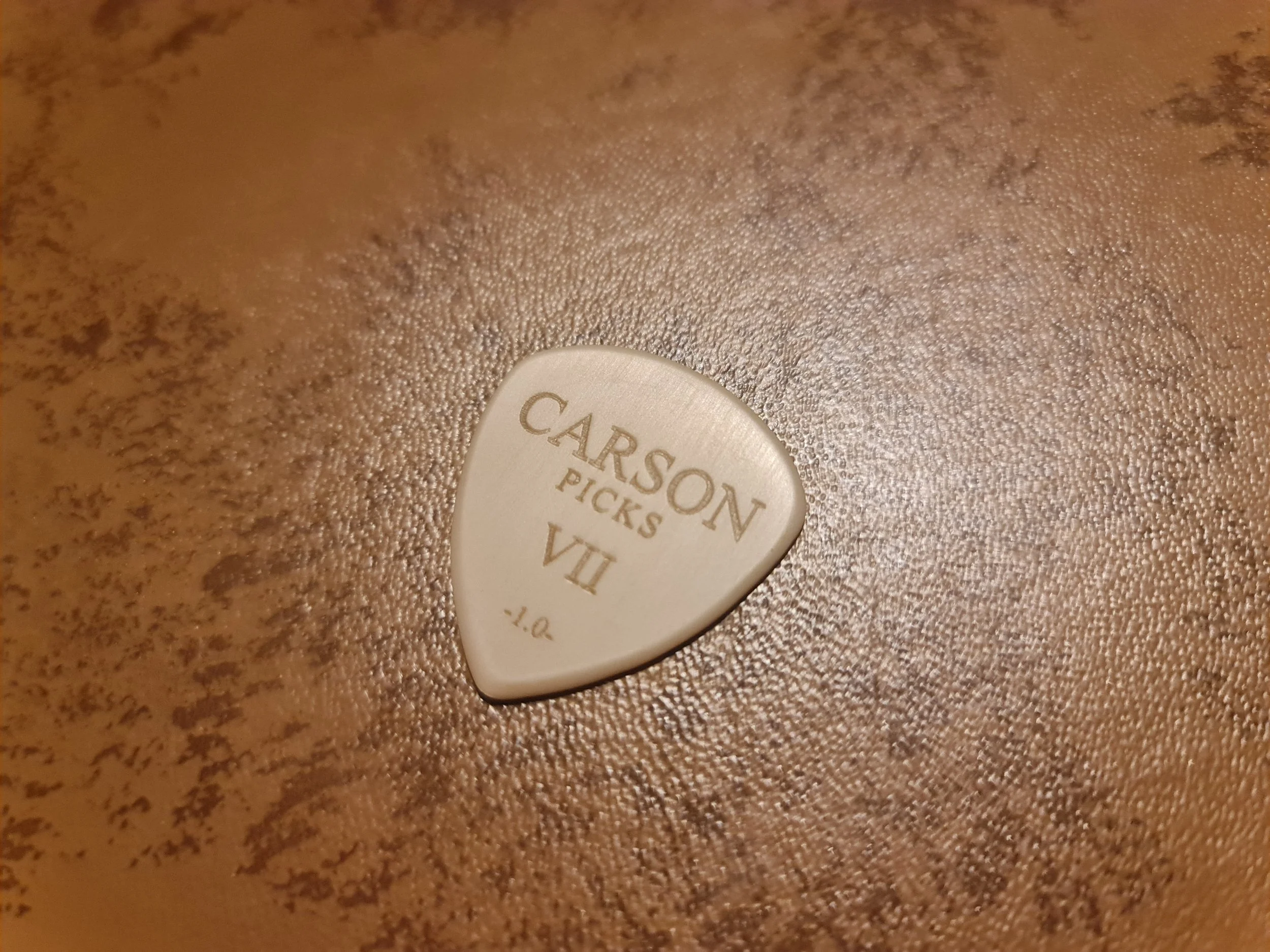 Close-up of a PEEK guitar pick with the words 'CARSON PICKS' engraved on it, placed on a textured brown surface of a guitar case.