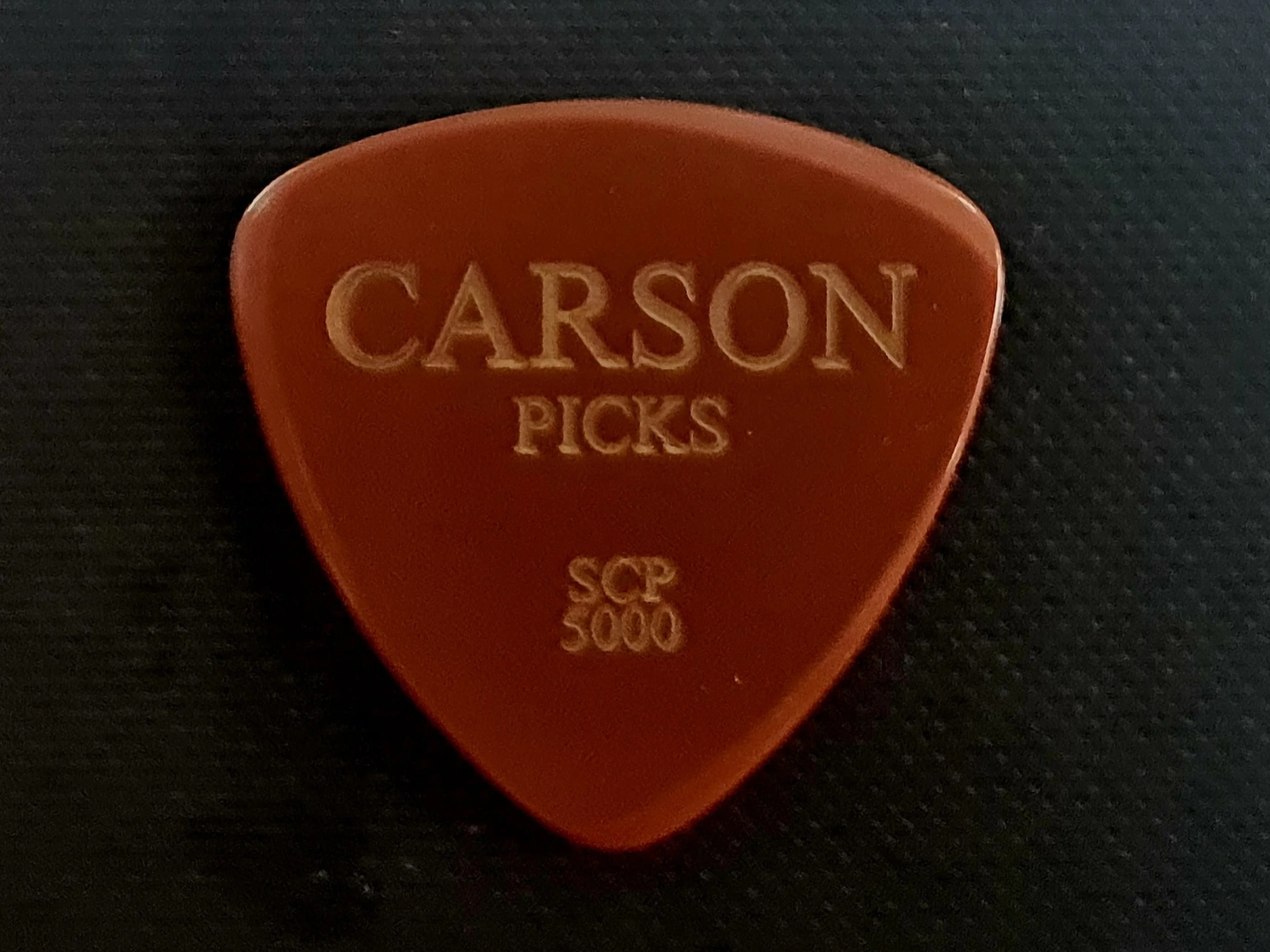 Close-up of a Vespel SCP-5000 guitar pick with the words "CARSON PICKS" engraved on it, resting on a dark textured surface. Made by Carson Picks