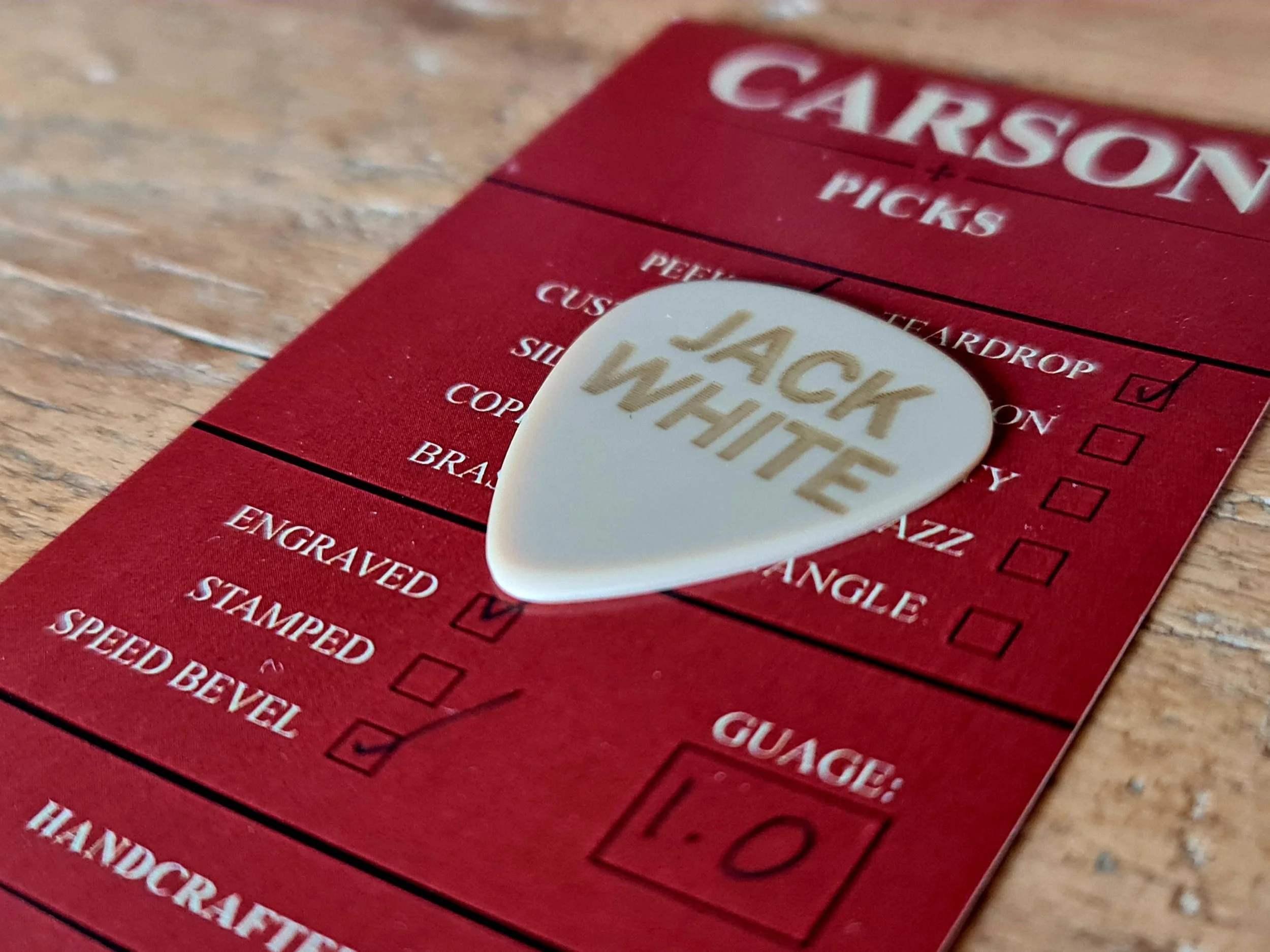 Close-up of a red packaging insert with a white PEEK guitar pick on top that has 'JACK WHITE' engraved on it.