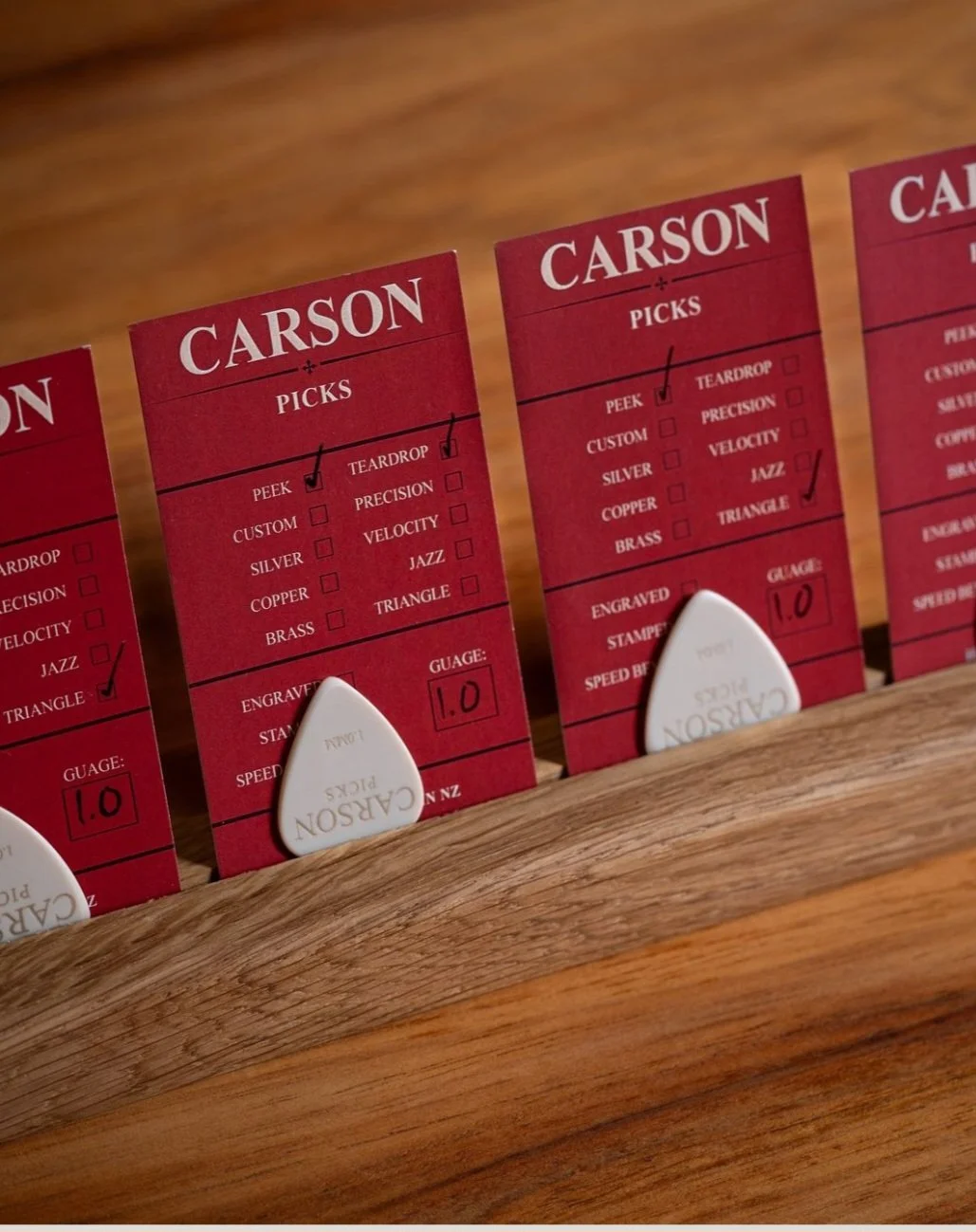 Three red cardboard packaging slips labeled "Carson Picks" with checkmarks, sitting on a wooden surface with white PEEK guitar picks partially visible underneath.
