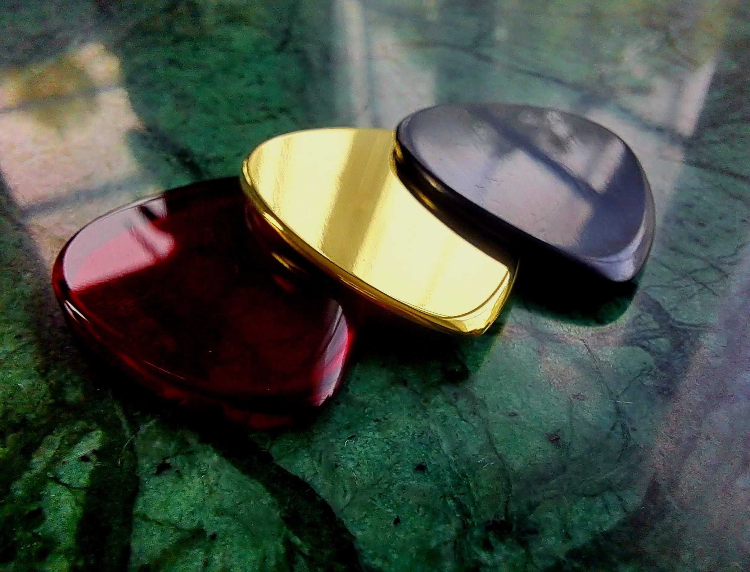 Three guitar picks made by Carson Picks. Two in acrylic, red and and black and one in high polish brass. 3mm thick