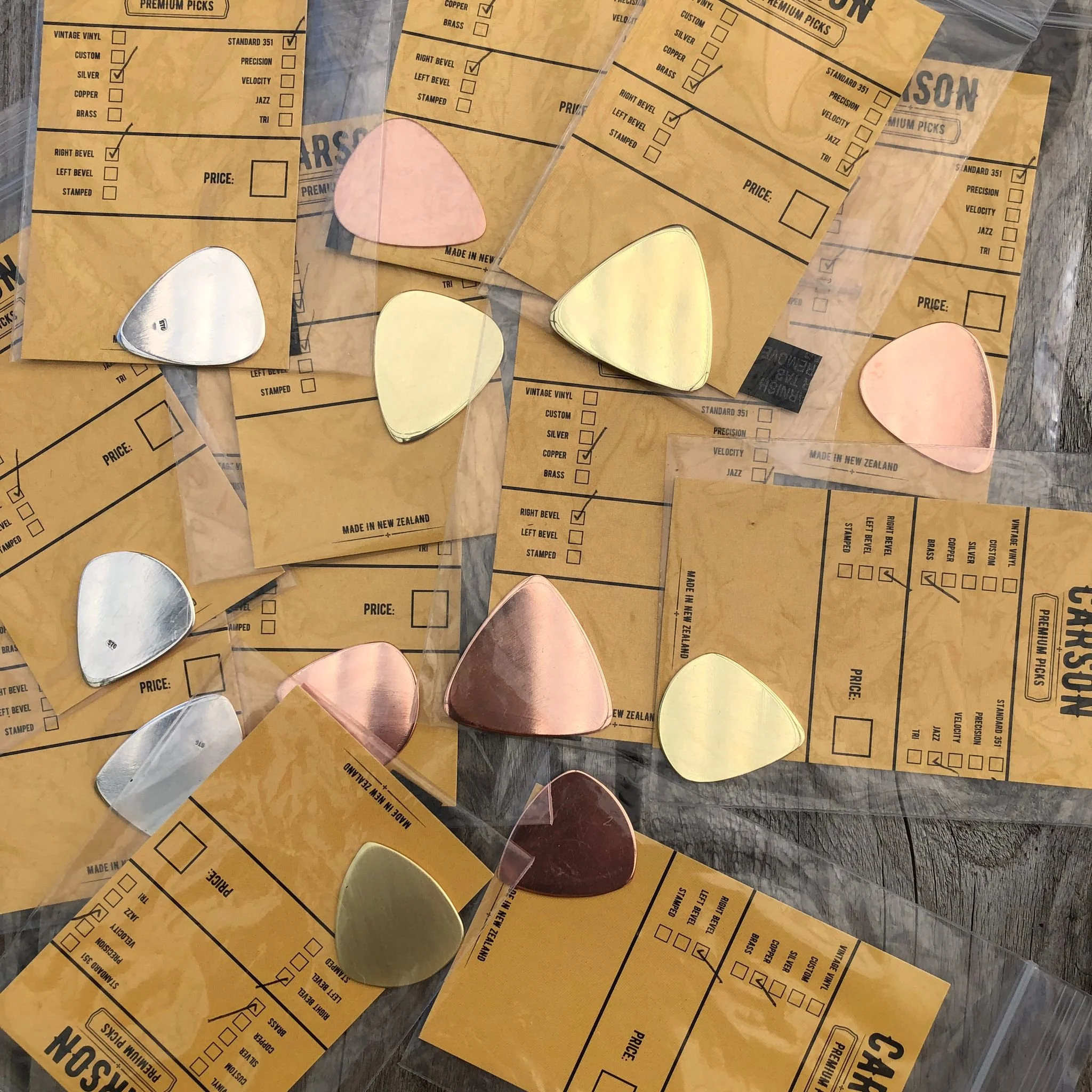 Several guitar picks by Carson Picks in different metals such as silver brass and copper placed on yellow shopping bags with checkboxes for selecting guitar pick options.