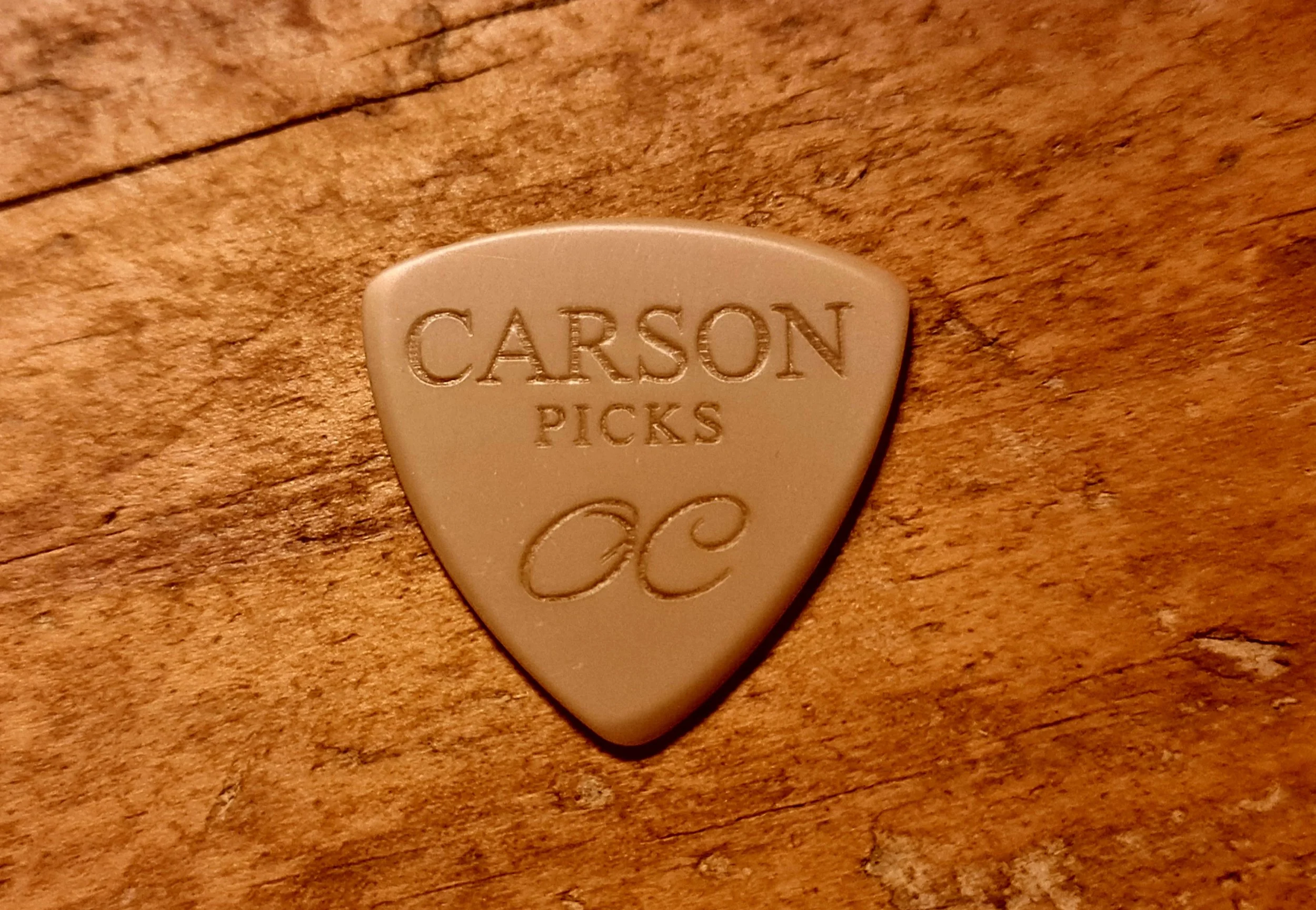A beige PEEK guitar pick with the text 'Carson Picks' and a signature, placed on a wooden surface.