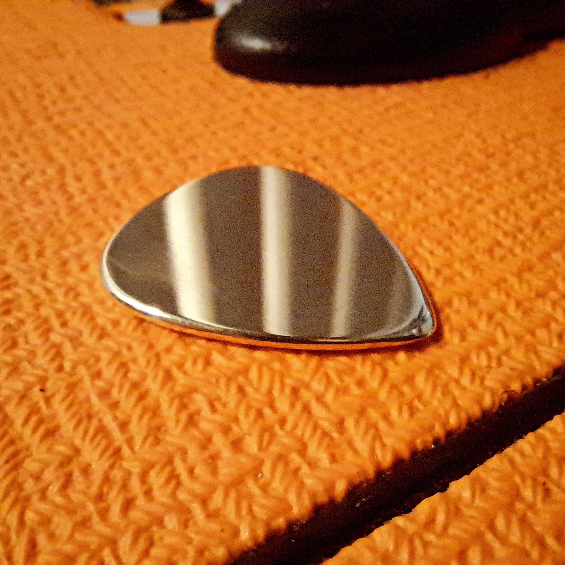 Close-up image of a Carson Picks brass pick in a high polish, laying on an orange amp.