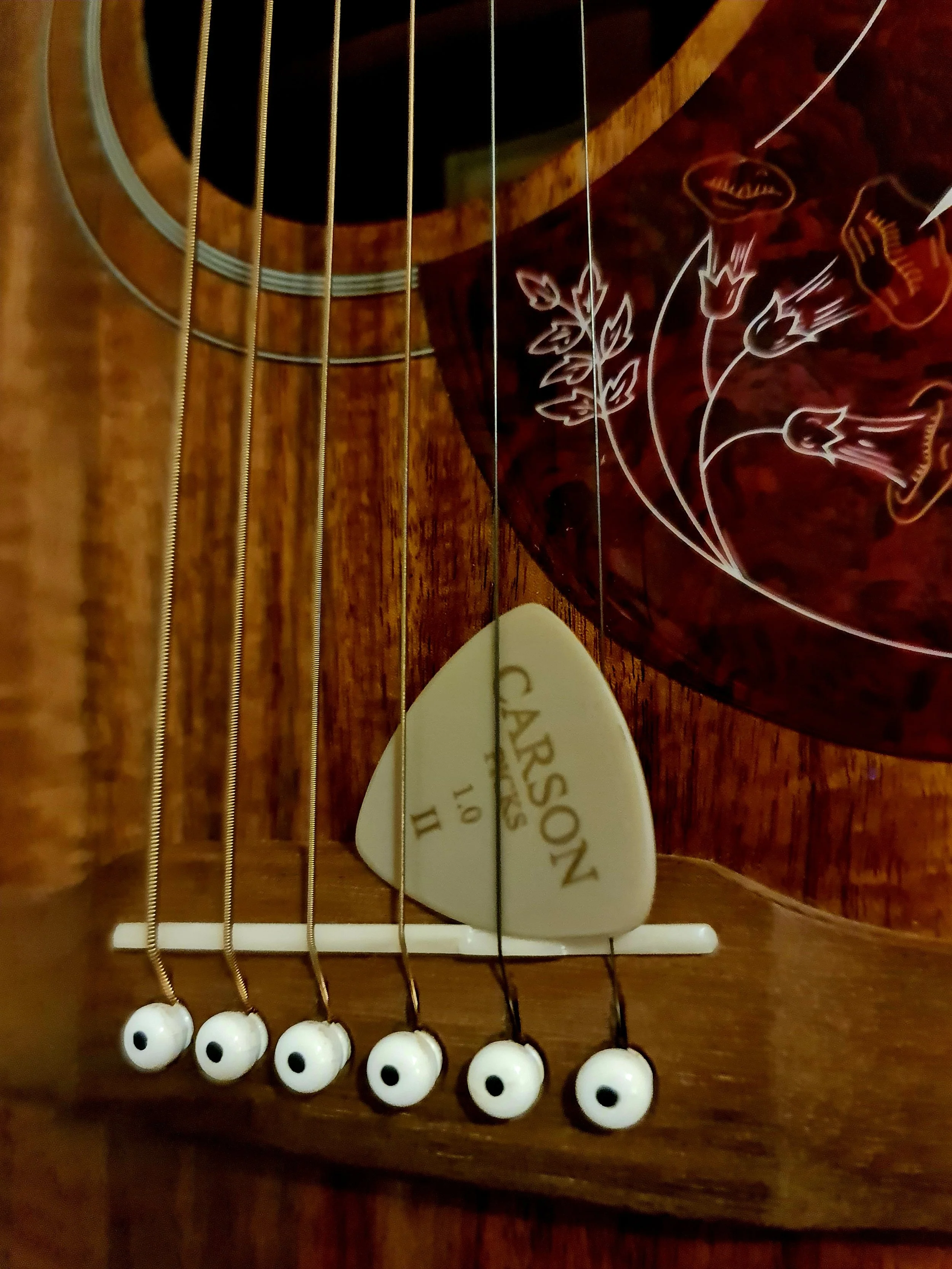 Close up of a Carson Picks PEEK pick tucked underneath the strings, near the bridge of an acoustic guitar.