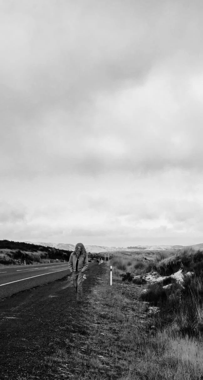 A person walking along a roadside in a rural landscape with cloudy sky and rolling hills in the distance, captured in black and white.