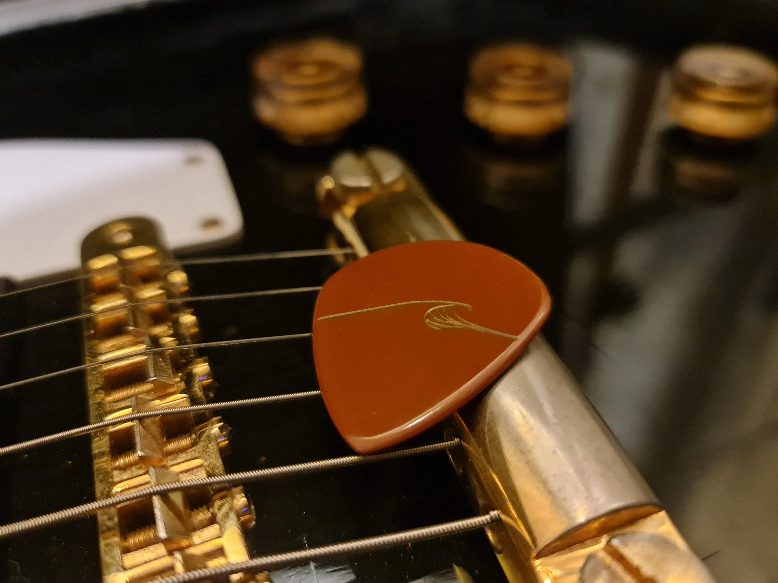 Close-up of a guitar's bridge, strings, and volume controls, with a Carson Picks Vespel SP-1 pick resting on top, with Eddie Vedder's wave logo engraved on it.