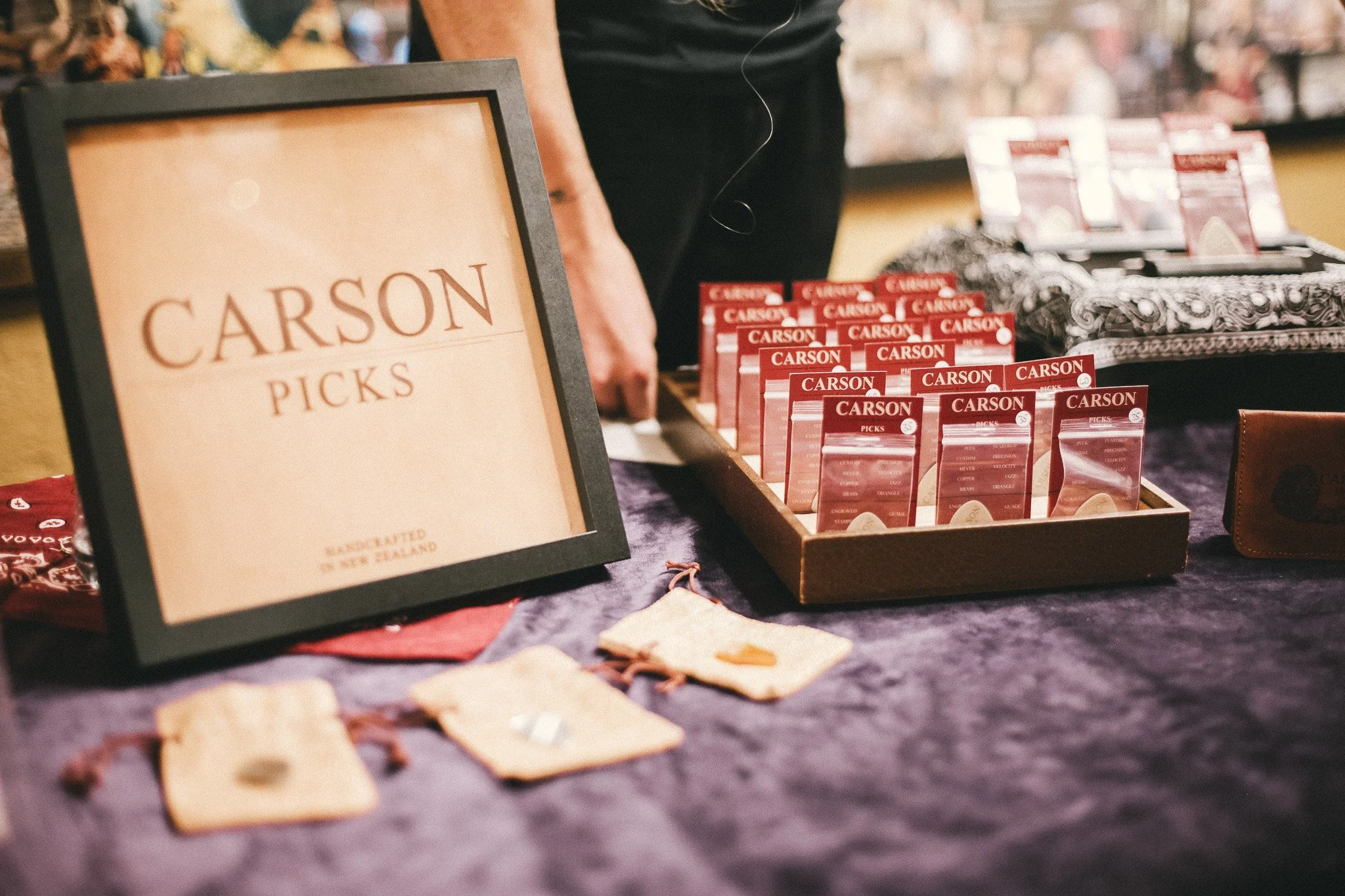 A booth with a sign that reads 'Carson Picks', various red packing inserts with white text, and a variety of PEEK, sterling silver and stone guitar picks on a dark purple cloth. Titirangi market, lopdell