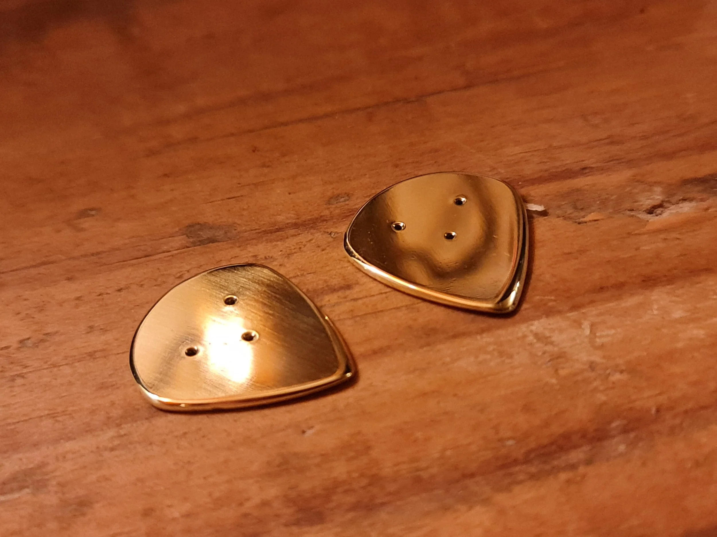 Close-up of two brass high polish jazz picks by Carson Picks with three small grip holes, resting on a wooden surface.